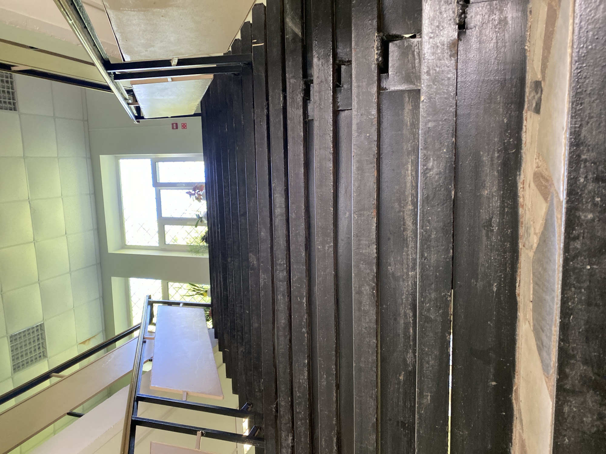 The image presents an interior view of a building's stairwell, captured from an elevated position looking down a set of stairs.
On the right side of the frame, a dark, open-riser staircase descends. The stair treads and the vertical support beams appear to be made of a dark material, possibly painted metal or heavily painted wood, exhibiting some texture and wear. A handrail system runs alongside the stairs, featuring a metallic top bar and lighter, possibly wooden or paneled, sections underneath.
To the left of the stairwell, a bright interior space is visible, likely a hallway or room. It features a ceiling composed of light-colored square tiles, with one square ventilation or lighting fixture partially visible. Two large windows dominate this wall, letting in significant natural light, indicating it is daytime. These windows are composed of multiple panes, forming a distinct grid pattern. A small green plant is placed on the sill of the lower window.
On the wall, positioned between the ceiling and the upper window, two small red square signs with white icons are visible. One clearly depicts an emergency exit symbol of a running person. The other is a red square featuring a white circular symbol, whose precise meaning is not fully discernible but appears to be safety-related. No explicit readable text is present on these signs.
The abundant natural light suggests it is daytime. The architectural elements, such as the ceiling tiles and the style of the stairs, imply the setting is likely an institutional or commercial building, possibly an older structure. There are no people visible, and no discernible activity is taking place.