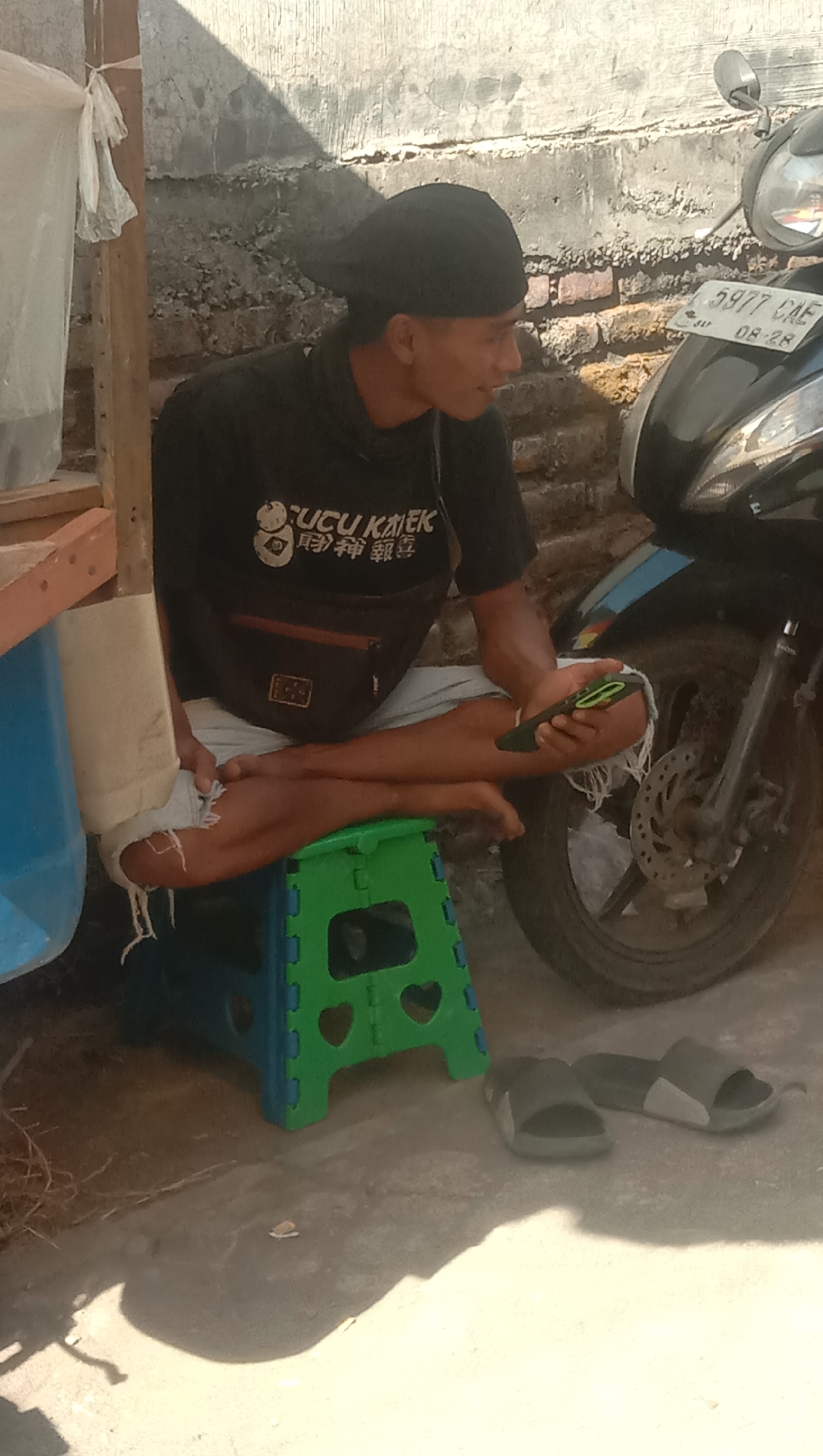 A man is sitting cross-legged on a green and blue plastic stool outdoors, next to a brick wall. He is wearing a black t-shirt with white text and graphics, a black baseball cap, and light-colored shorts. He is holding a dark-colored smartphone in his right hand and appears to be looking at it or speaking. A black motorcycle is parked to his right, with its license plate visible reading "5977 CAE 08-28". In the foreground, a pair of dark gray slides are on the ground. To the left of the man, a wooden post supports a white plastic bag. The scene appears to be in a warm, sunny environment. This setting is consistent with a residential or roadside area in Karanganyar, Indonesia.