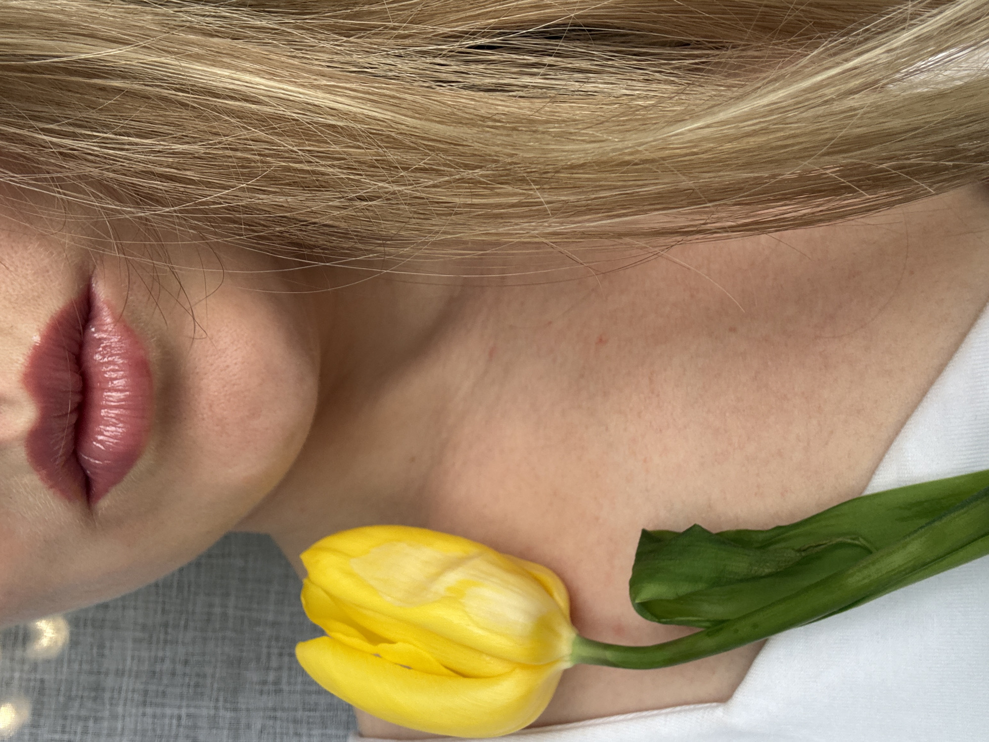 The image shows a person's head and upper chest, lying on a light gray surface. The person has blonde hair, and their lips are prominent with a glossy mauve-pink lipstick. A yellow tulip lies near their neck. A portion of a white garment is visible on the right side of the image. The location is not discernible from the information available in the image.