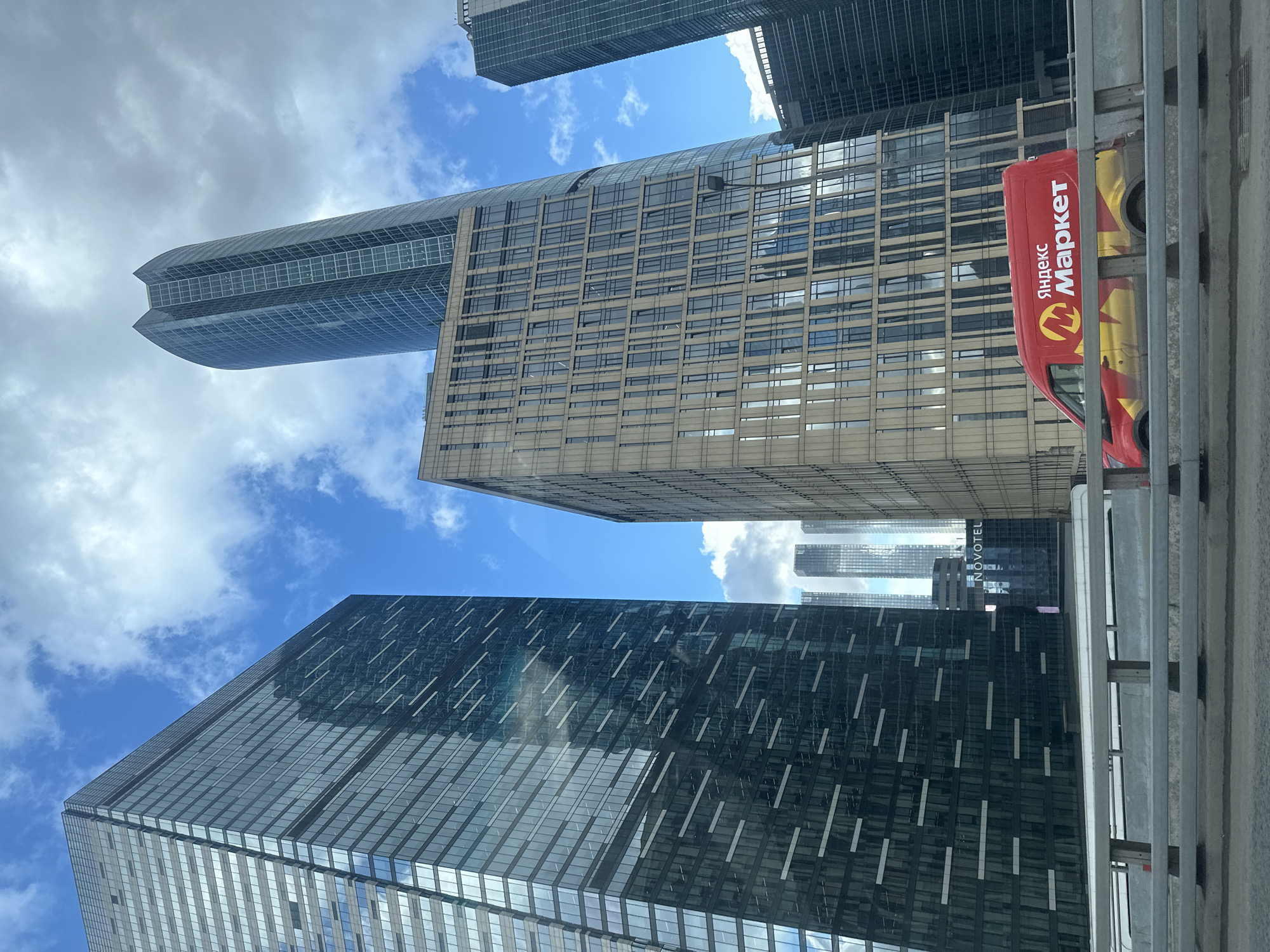 The image shows a view of the Moscow International Business Center ("Moscow City"), Moscow, Russia, from a low angle. Several modern skyscrapers with glass facades dominate the frame. The sky is visible between the buildings, with a mix of blue sky and clouds. A red and yellow delivery van with the Cyrillic text "Яндекс Маркет" (Yandex Market) is partially visible on the right side. A metal barrier or railing is at the bottom of the image, suggesting the photo was taken from a road or walkway.