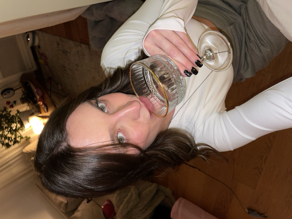 A woman with dark brown hair and green eyes poses close to the camera, holding two glasses. She has dark nails and is wearing a white long-sleeved shirt. She's sticking her tongue out, touching the rim of the glass closer to the camera. The background shows a room with a floor lamp and a table, suggesting a casual indoor setting.