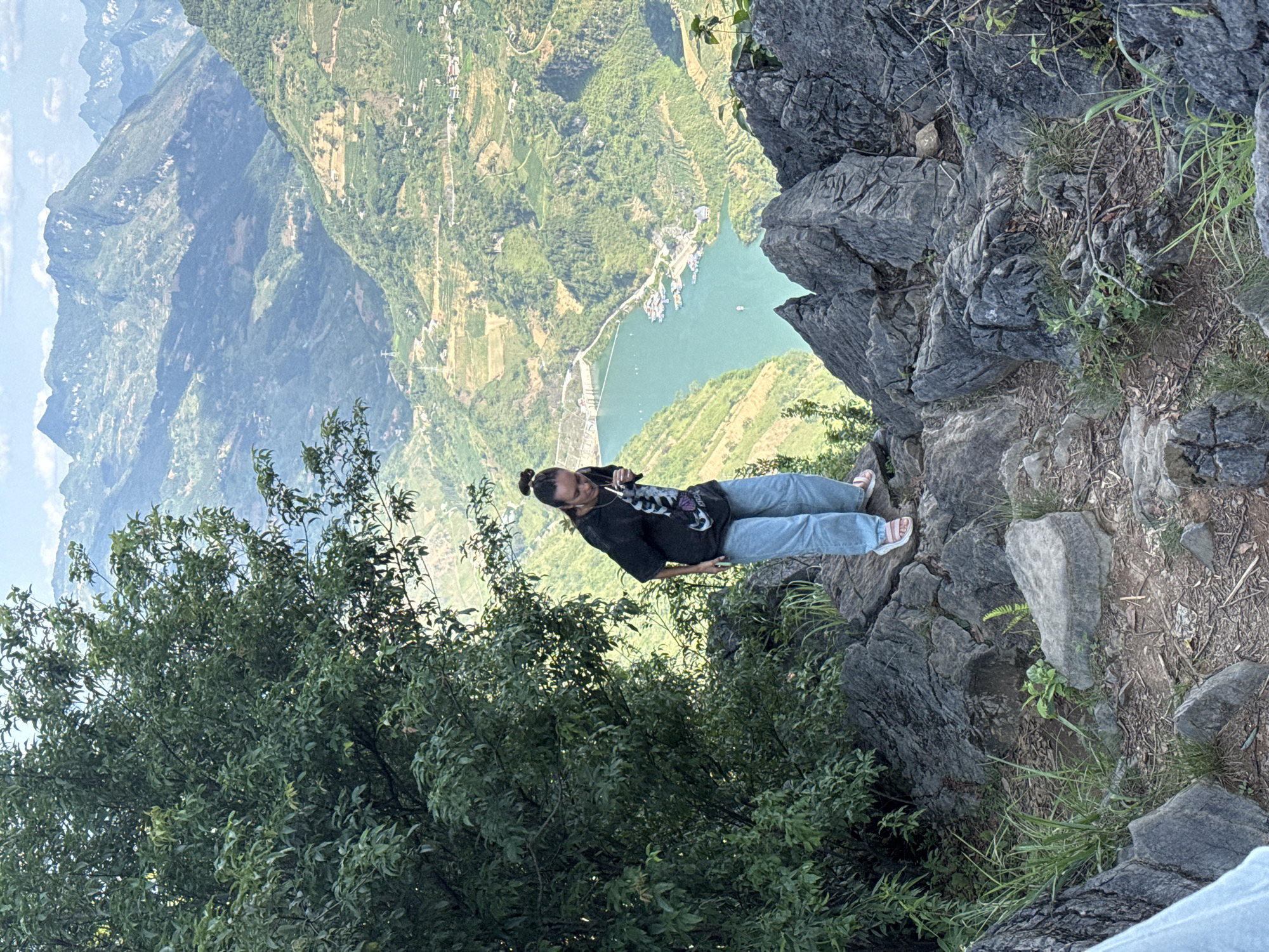 A woman sits on the rocky edge of a cliff. She is wearing a dark t-shirt, light blue jeans, and sandals. She appears to be looking at something in her hand. Lush green trees and vegetation are in the foreground, partly obscuring the view. A vast mountainous landscape stretches out behind her, with terraced fields and a body of water visible. This landscape indicates the location is likely within the Đồng Văn Commune, Vietnam. The cliffs appear rocky and naturally formed, indicative of a mountainous environment.