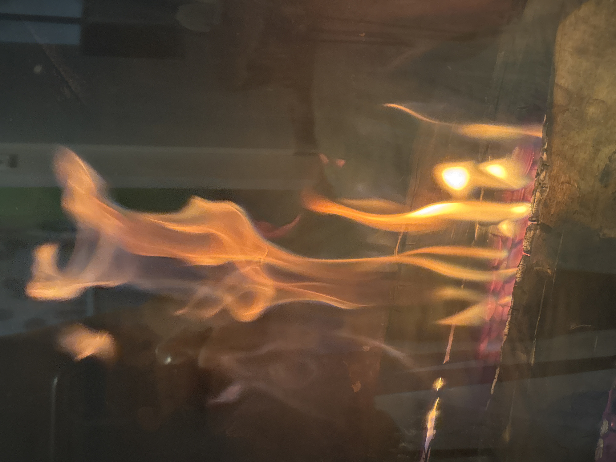 The image shows a close-up view of a fireplace with burning logs. The flames are orange and yellow, with some hints of pink and blue at the base. The logs are dark brown and appear to be made of wood. The fireplace is likely made of brick or stone, as suggested by the texture and color of the materials visible. There are reflections in the glass of the fireplace, which obscure some of the details of the surrounding environment. It is not possible to determine the specific location within Zvenigorod, Russia based solely on this image.