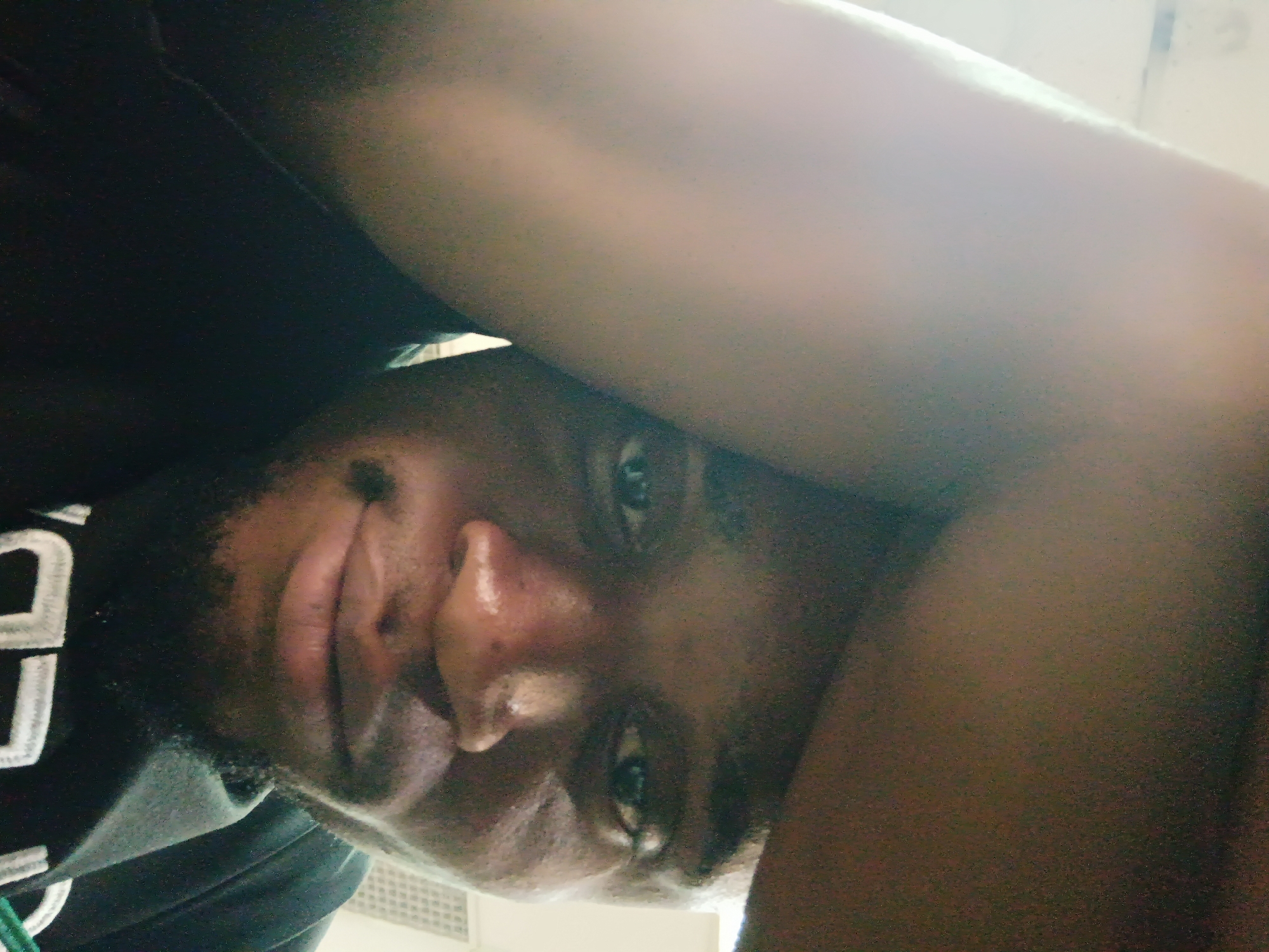 This close-up, tilted portrait features a young adult male of African descent, looking directly at the camera with a neutral to slightly pensive expression. The image is captured from a low angle, focusing on his face, which appears to have a short beard and mustache. He is wearing a dark top, possibly a t-shirt or hoodie, with partial white lettering that reads "CD" or similar characters visible on the left side of his chest.
The activity appears to be posing for a close-up photograph, likely a selfie. Strong lighting from the upper right creates a significant glare across the top part of the image and illuminates his face, highlighting a sheen on his skin, which could suggest sweat or a humid environment. This harsh lighting also casts pronounced shadows, emphasizing the contours of his face.
The setting is an indistinct indoor environment, characterized by dark surfaces dominating the upper left and lighter, possibly off-white, surfaces on the right and bottom. A faint grid-like pattern, resembling a vent or grill, can be discerned in the lower right portion of the background, providing a subtle location cue for an indoor space. The time of day and weather are not discernible due to the indoor setting and the strong, possibly artificial, light source. The location context for this image is Kakuri Gwari, Nigeria.