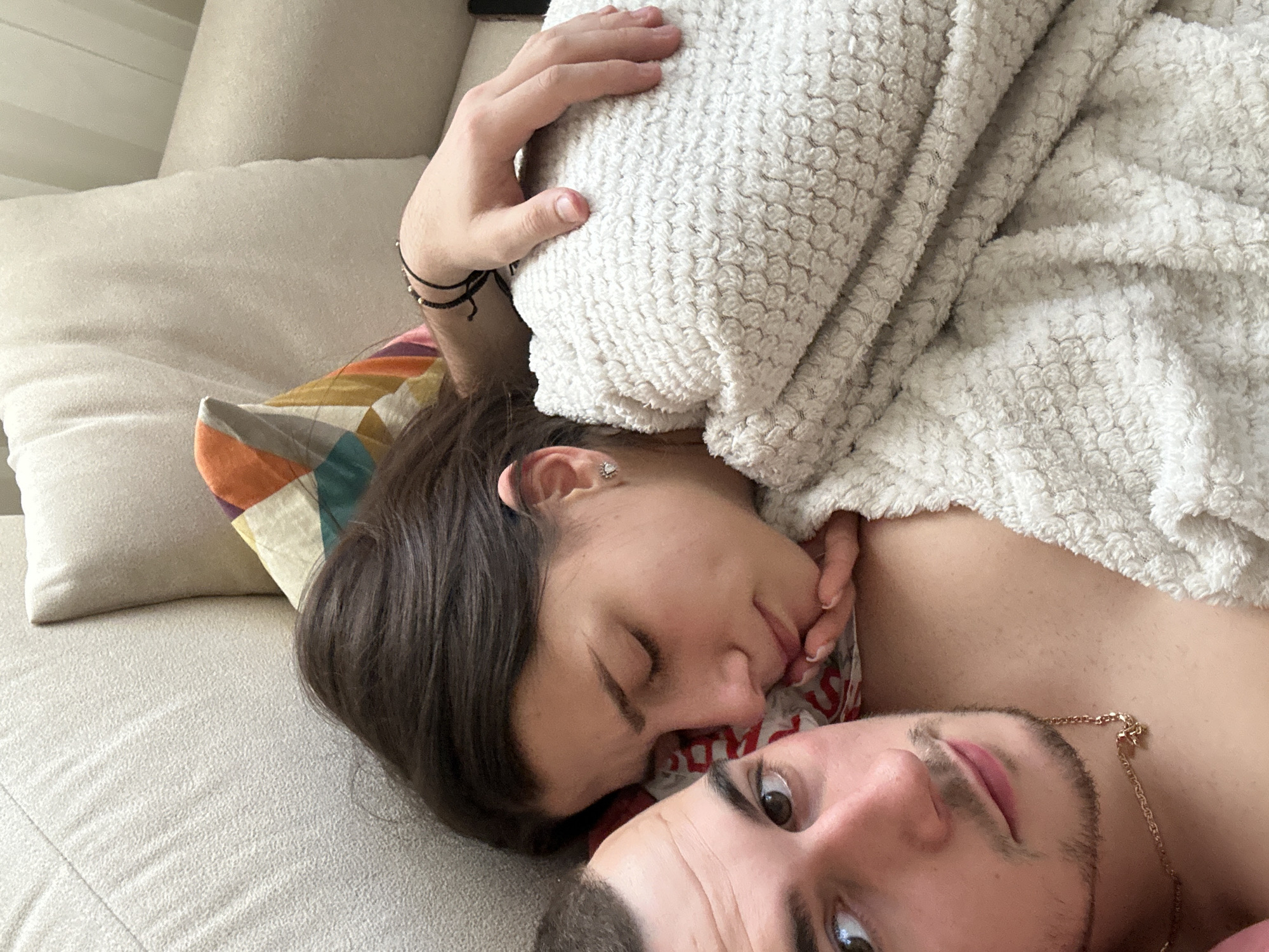 A couple lies on a beige sofa with throw pillows. The woman's head rests on a pillow with a geometric pattern, covered with a patterned blanket. Her right hand is above her head. The man's face is visible at the bottom of the frame. The location is unknown and there is no information to indicate Plovdiv, Bulgaria.