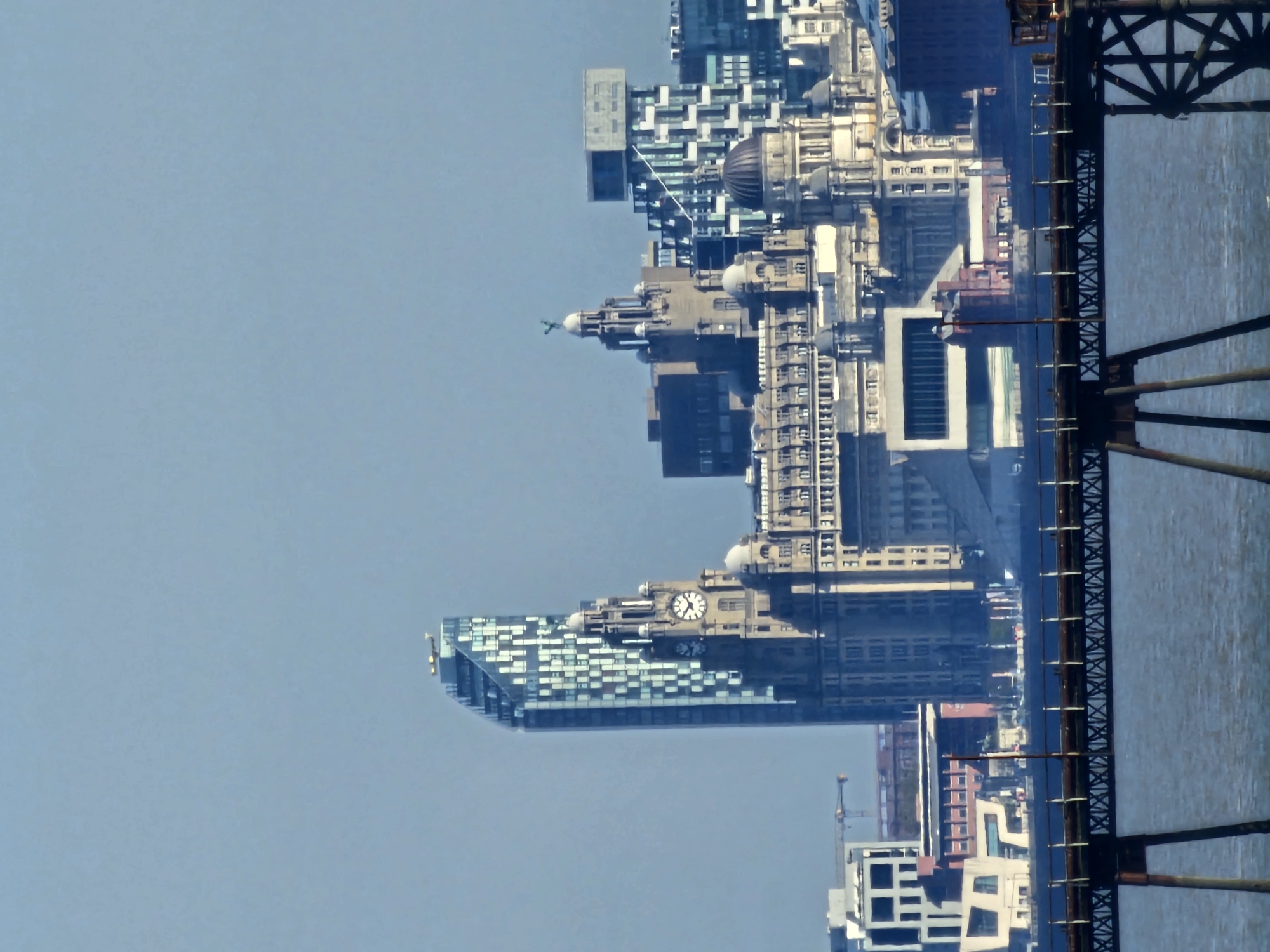 An outdoor, daytime image is oriented vertically, showcasing a dense cityscape on the left, a body of water, and a prominent bridge structure on the right, all beneath a clear blue sky that occupies the left portion of the frame.
The cityscape, likely the Liverpool waterfront viewed from New Ferry, United Kingdom, features a striking blend of historic and contemporary architecture. Dominating the upper-mid section are grand, ornate buildings with classical facades, domes, and spires, including one resembling the Royal Liver Building with its distinctive clock face and a bird-like figure atop a spire. Another building displays a clear clock with Roman numerals. Interspersed and adjacent to these older structures are modern, glass-fronted skyscrapers with geometric patterns, reflecting the bright sky. Some areas within the city show scaffolding or crane structures, suggesting ongoing development or maintenance.
To the right, a calm body of water, presumably the River Mersey, exhibits subtle ripples on its surface. Partially obscuring the water and parts of the distant city is a robust, dark-colored metal bridge or pier structure. This industrial-looking framework features intricate latticework, cross-bracing, and vertical supports, extending into the foreground on the right.
No people or specific activities are discernible in the scene. The weather appears bright and clear, consistent with daytime conditions. The exact time displayed on the clocks is not clearly legible. The overall impression is a vibrant urban panorama juxtaposed with a significant waterway and industrial infrastructure.
