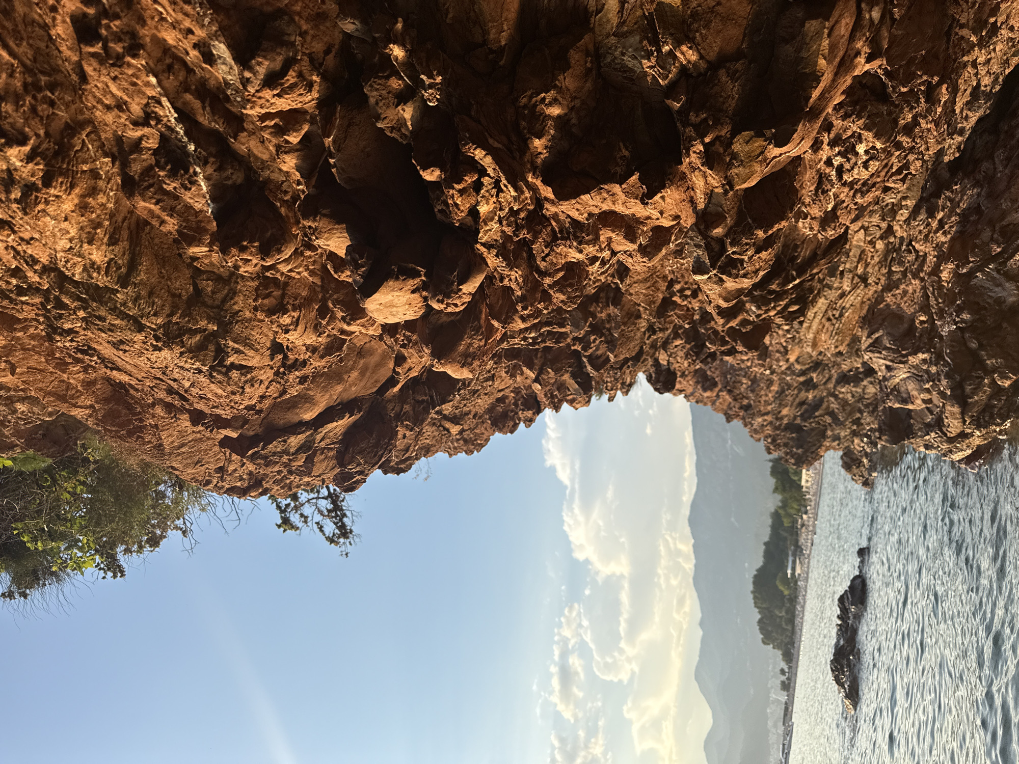 The image depicts a view from within a rocky overhang, looking outwards towards a bright sky and a body of water. The overhang itself is composed of rough, reddish-brown rock formations, with some vegetation visible on the left side.
Beyond the rocks, the scene opens up to a wide expanse of light blue sky with scattered white clouds. The sun is visible, casting a warm glow and creating subtle rays of light. In the distance, hazy mountains rise, partially obscured by the clouds and atmospheric conditions.
To the right of the sky, a glistening body of water stretches out, appearing to be the sea or a large lake. The water's surface is textured with ripples and reflects the light, suggesting it is calm. A few dark rocks are submerged or partially visible within the water. Further along the shoreline, a distant landscape features trees and what appears to be a small settlement or buildings, hinting at a coastal or lakeside location.
The orientation of the image is such that the rocky overhang is at the top, and the sky and water are below, as if looking up and out from a cave or alcove. The lighting suggests it is daytime, likely late afternoon or early evening, given the warm hues and the angle of the sun. The overall impression is one of natural beauty and tranquility, with the ruggedness of the rocks contrasting with the open expanse of the sky and water. No people, objects, or text are clearly discernible in the frame.