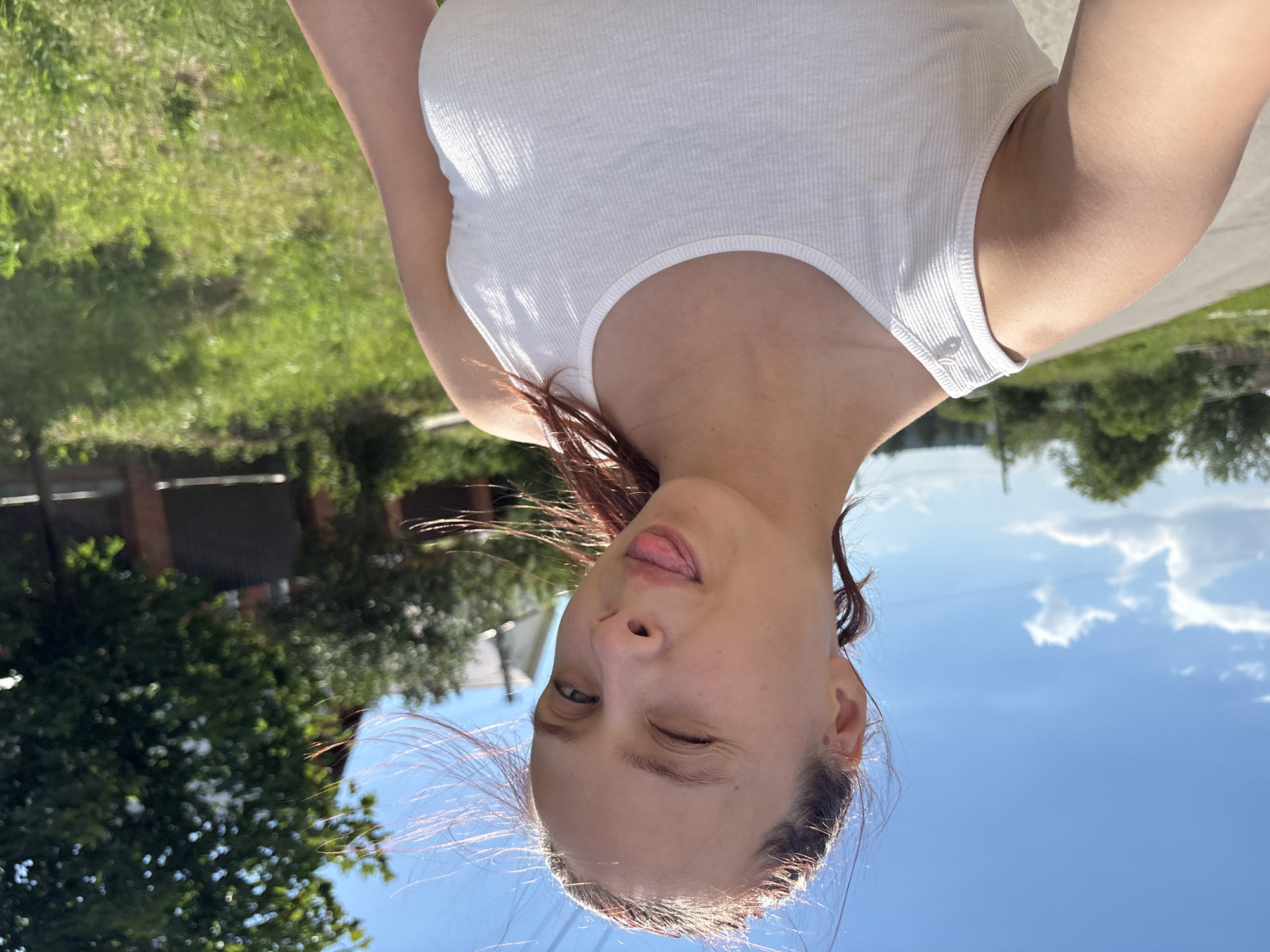 A young woman with red hair tied back in a ponytail is taking a selfie outdoors on a sunny day. She is wearing a white ribbed tank top and is winking with her tongue sticking out. The background shows a rural landscape with a paved road, green grass, trees, and a brick fence. In the distance, a house with a red roof is visible. The image appears to have been taken in a location resembling the outskirts of a city or town, potentially in a rural area of Russia given the architectural style of the house and the vegetation.