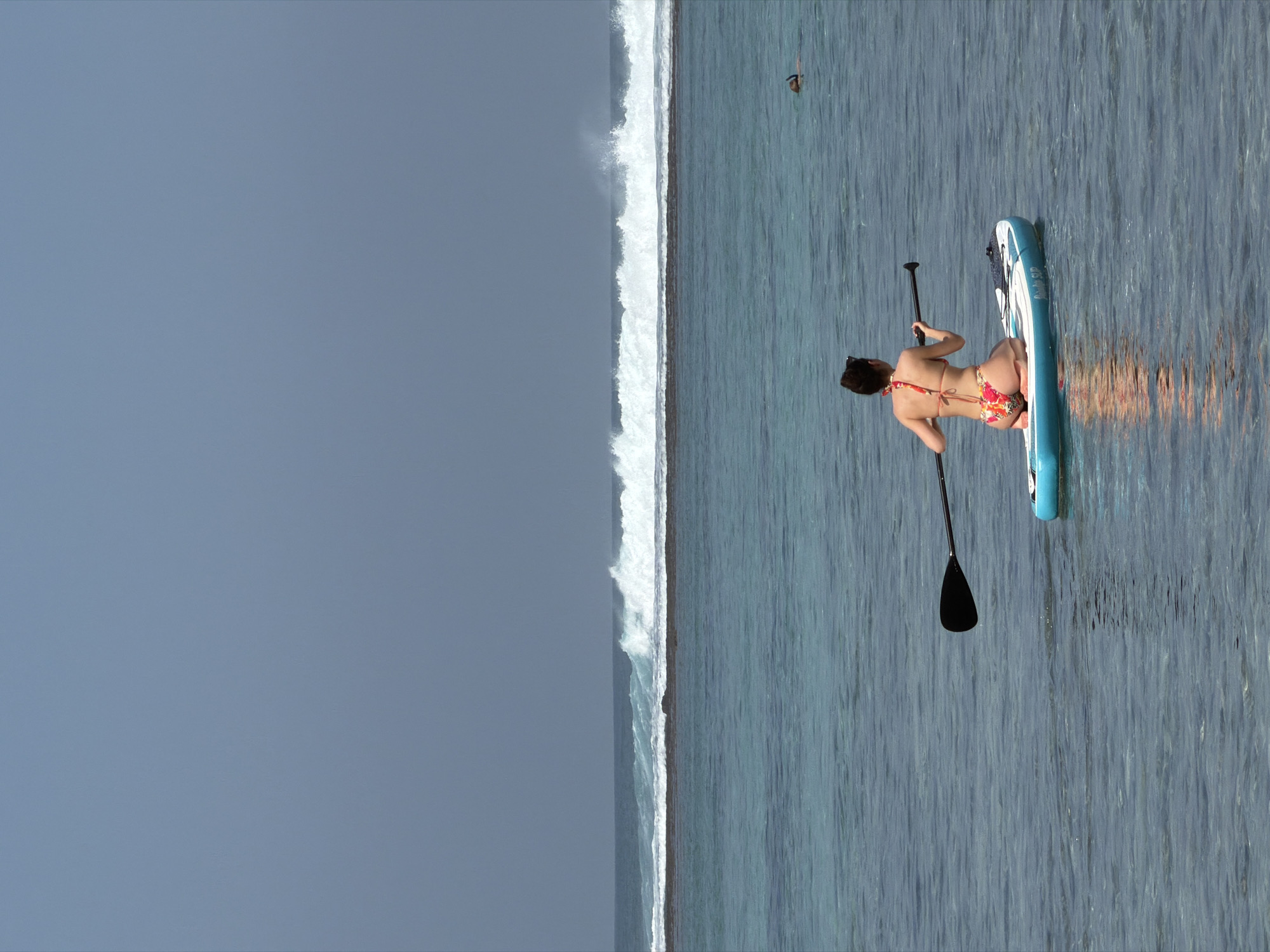 An overhead shot captures a woman paddleboarding in the ocean off the coast of Kuta Selatan, Indonesia. She is kneeling on a blue and white paddleboard, wearing a floral bikini, and holding a black paddle. The water is a clear turquoise, with gentle waves breaking in the distance against a reef. The sky is a pale blue, and a person is snorkeling further out in the water.