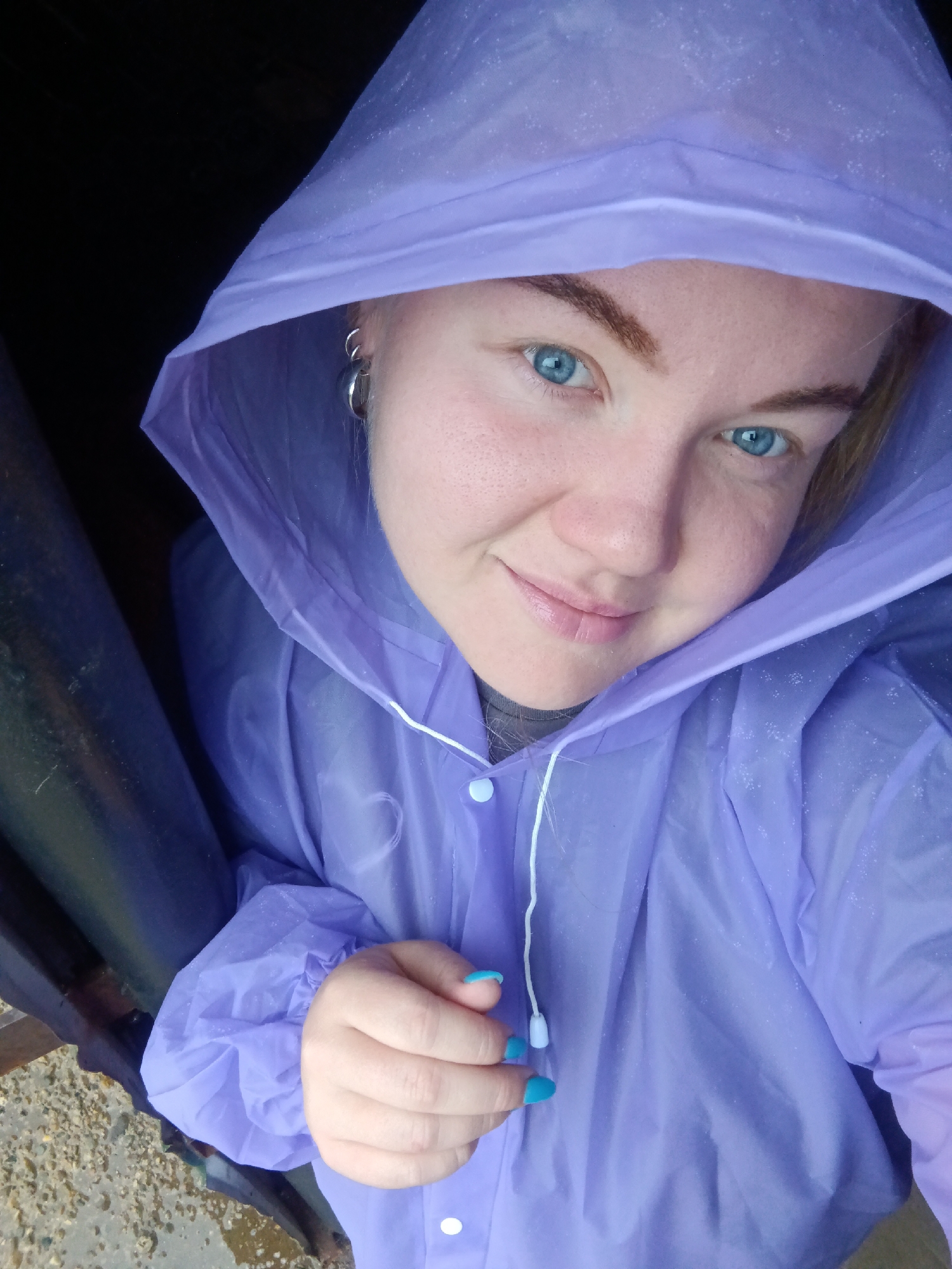 A young woman with bright blue eyes and blonde hair pulled back is looking at the camera from a close-up, high-angle perspective. She is wearing a light purple hooded raincoat. Her fingernails are painted a bright turquoise. The background is dark and out of focus, suggesting she may be under an overhang or in a doorway, with a hint of a dark cylindrical object to her left and wet, pebbly ground visible in the lower left corner. The overall impression is that she is posing for a selfie outdoors, possibly in the rain. No specific landmarks or details within the city of Foky, Russia, are discernible from the image.
