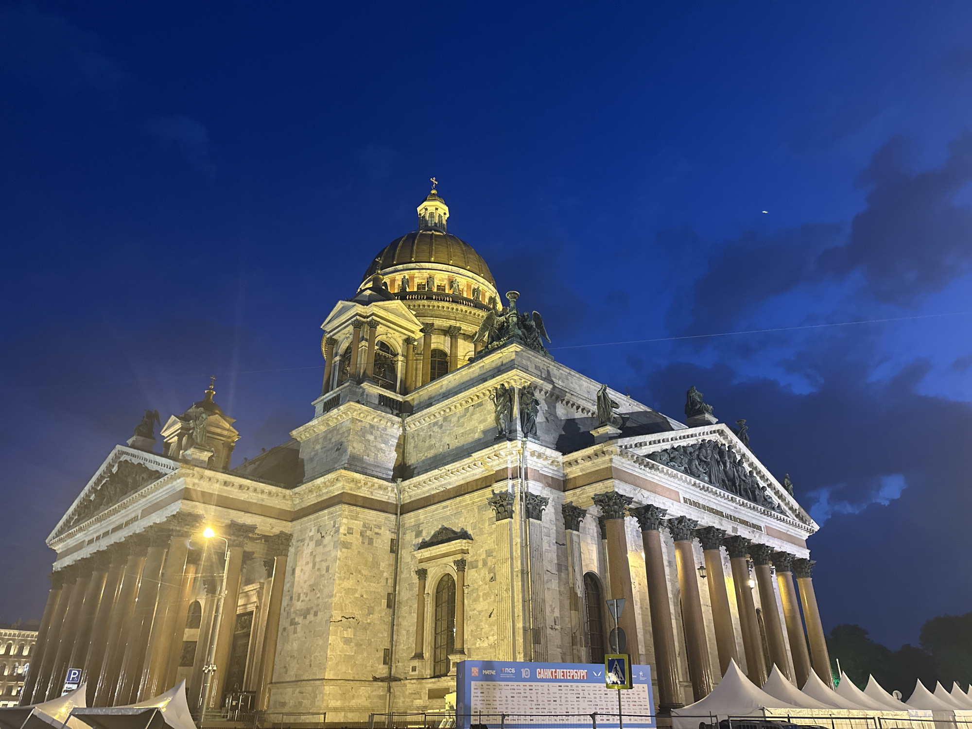 The image depicts St. Isaac's Cathedral in Saint Petersburg, Russia, at night. The cathedral is illuminated, and the sky is a deep blue with some clouds. A thin crescent moon is visible in the upper right. In the foreground, there is a sign with details about an event. There are some white tents visible in front of the cathedral.
