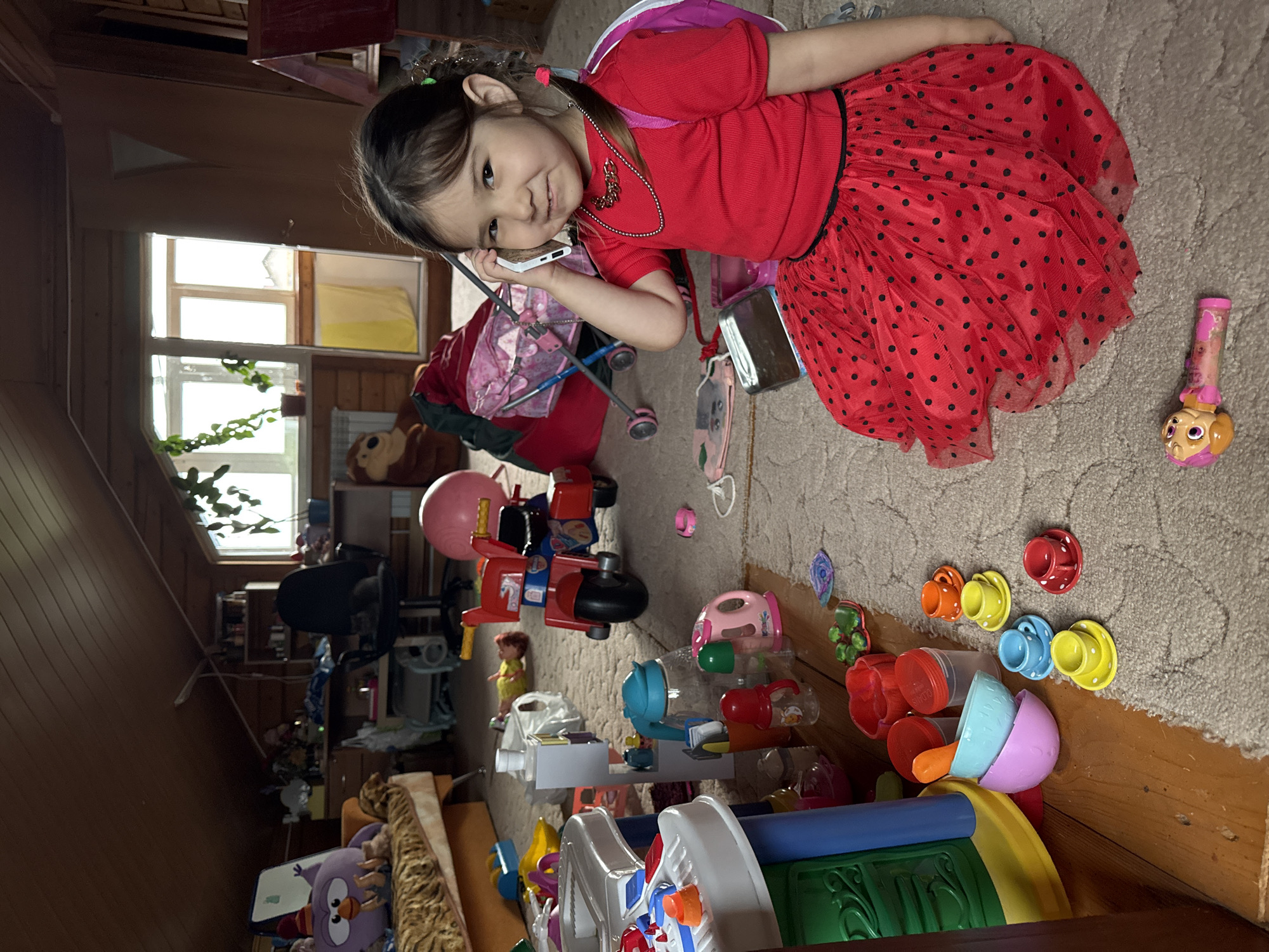 The image shows a young girl in a red polka dot dress and a red t-shirt, sitting on a carpeted floor, holding a white object to her ear as if on a phone. She has dark hair styled in pigtails. The room appears to be an attic or a room with a sloped ceiling, with wooden walls and ceiling beams visible. The floor is covered with a light-colored carpet. The room is cluttered with children's toys, including a red toy tricycle, a doll on the floor, a pink ball, a toy kitchen set, stacking cups, and various plastic containers and bottles. In the background, there is a couch, a desk with a chair, and windows. The overall impression is a child's play area in a residential setting. The image is not specific to Старое Михайловское, Russia, and there are no visible indicators of its location.