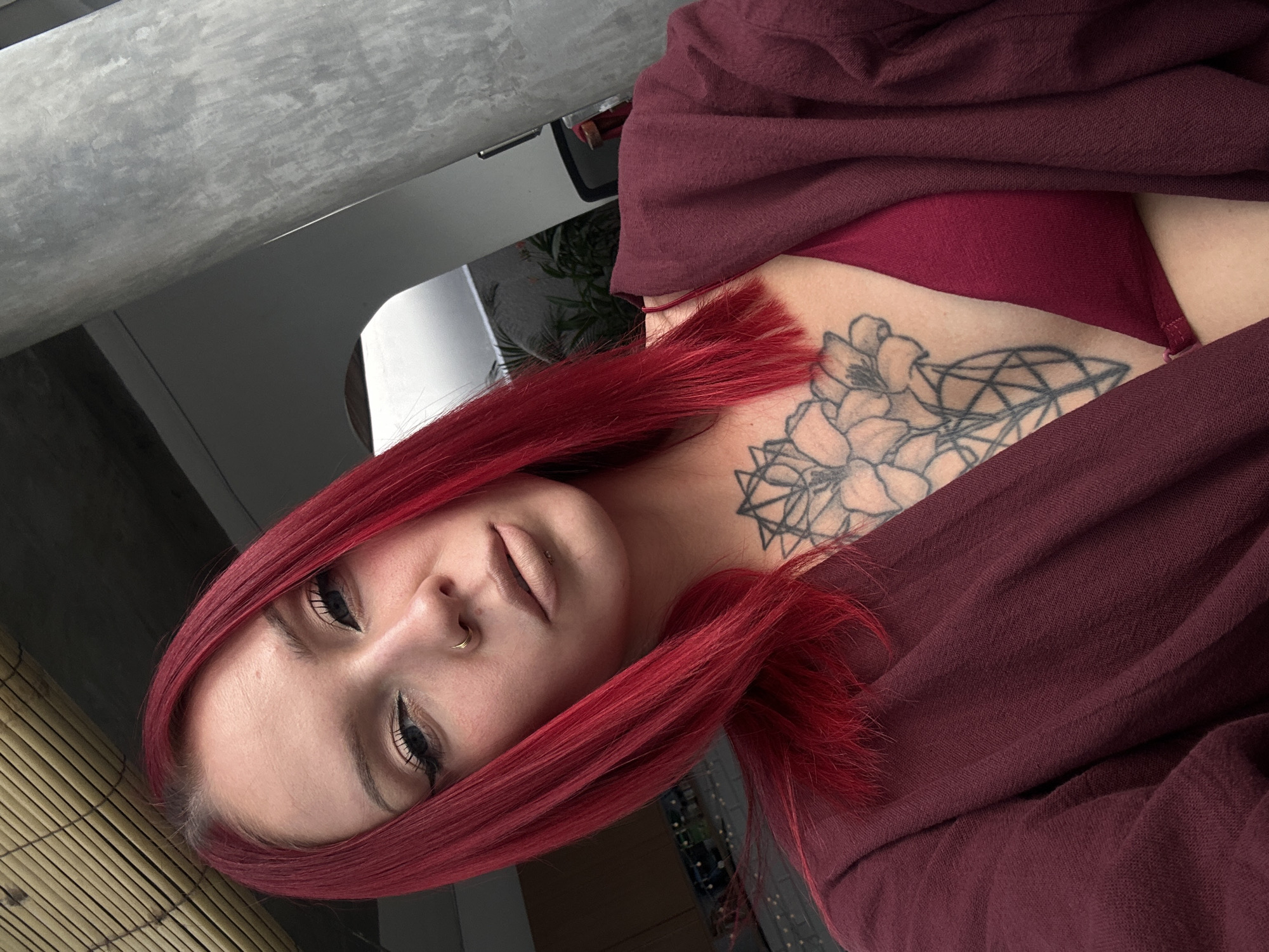 A woman with red hair is positioned looking towards the camera. She has a nose ring in her left nostril and is wearing makeup with eyeliner. A floral and geometric tattoo is visible on her upper chest/neck area. She is wearing a burgundy-colored garment, possibly a robe or shawl, over what appears to be a matching burgundy-colored bra or top. The background includes a window or glass panel with a blurred cityscape, a metal pole or beam, and a rolled-up bamboo screen or blind. The environment suggests an interior setting. There is no indication that the location is in North Kuta, Indonesia.