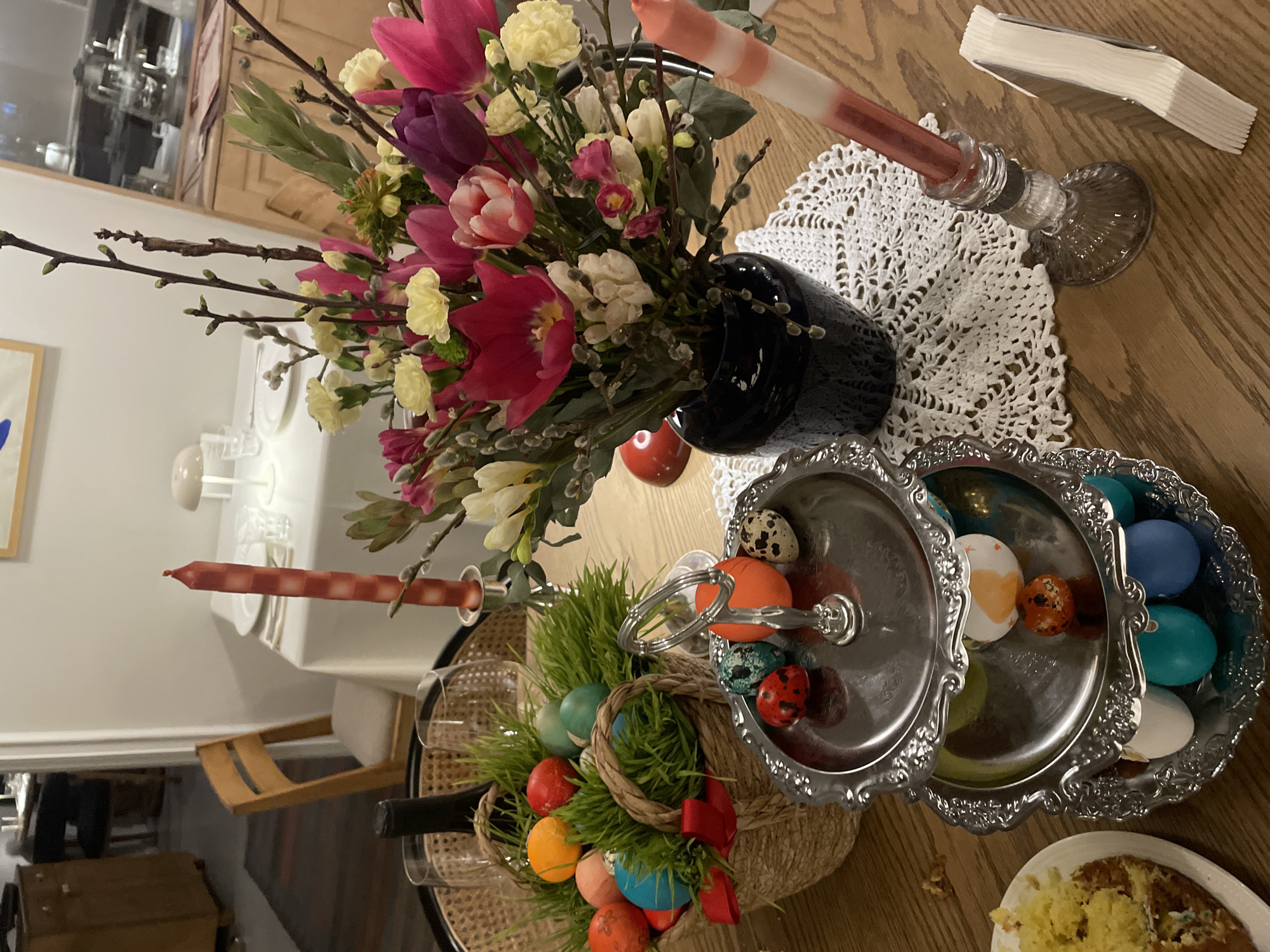 The image depicts a dining table laden with Easter decorations. A vase filled with tulips, pussy willow branches, and white carnations sits atop a white lace doily. A checkered candle in a glass candlestick holder is also present. An Easter basket filled with dyed eggs and faux grass sits beside a two-tiered silver serving dish containing more eggs. A plate of cake is also visible. The scene appears to be indoors. The location, Yerevan, Armenia, is not discernible from the image.