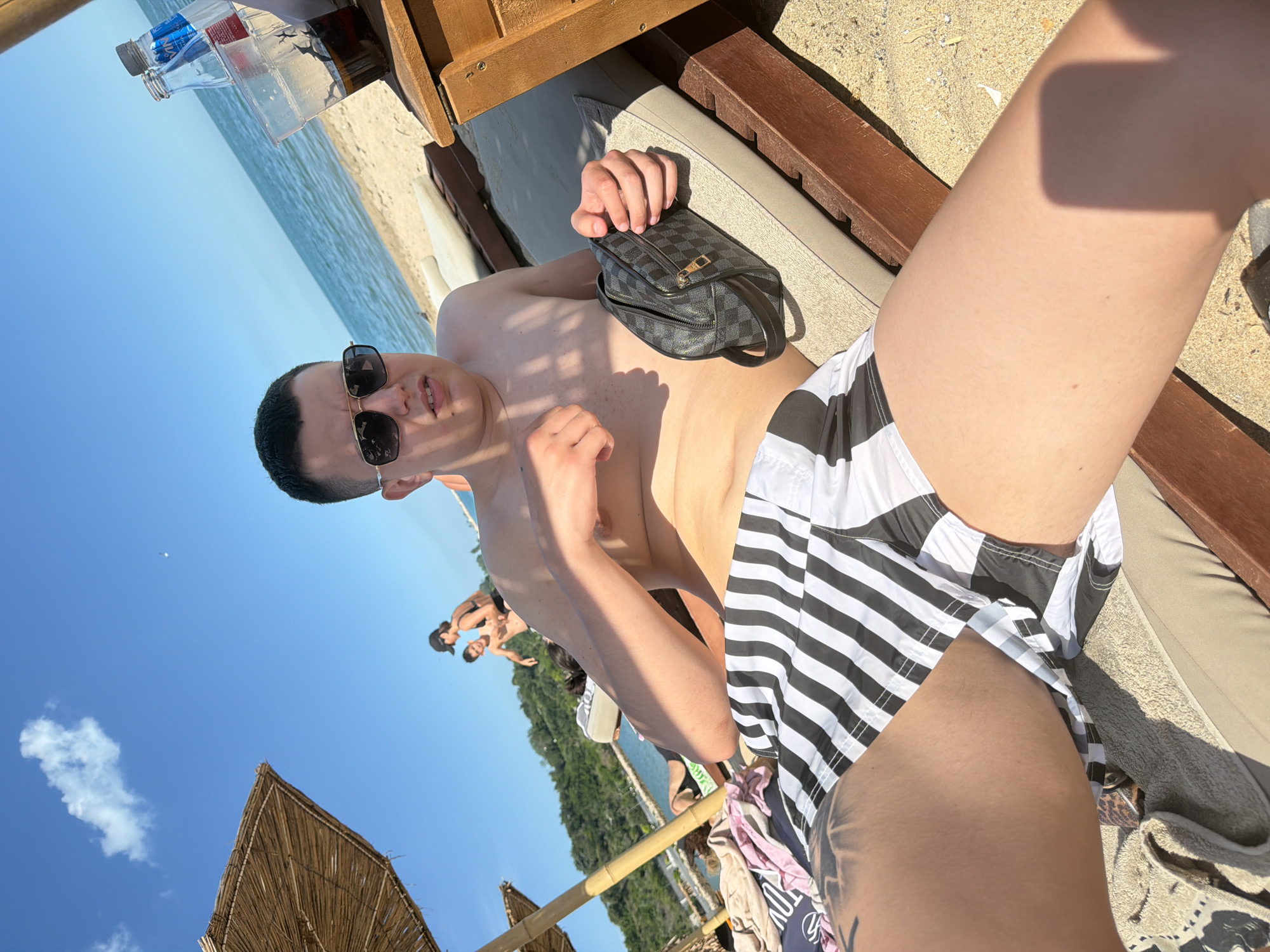 A young man with short dark hair and sunglasses is lying on a beach chair on a sandy beach. He is wearing black and white striped swim trunks. He is holding a dark patterned toiletry bag in his left hand. In the background, the sea meets the horizon under a clear blue sky with a few clouds. To the left of the man, a thatched umbrella provides shade. Further in the background, other beachgoers are visible near the water. A wooden side table with drinks is next to the man. The scene is consistent with a sunny day at a beach resort.