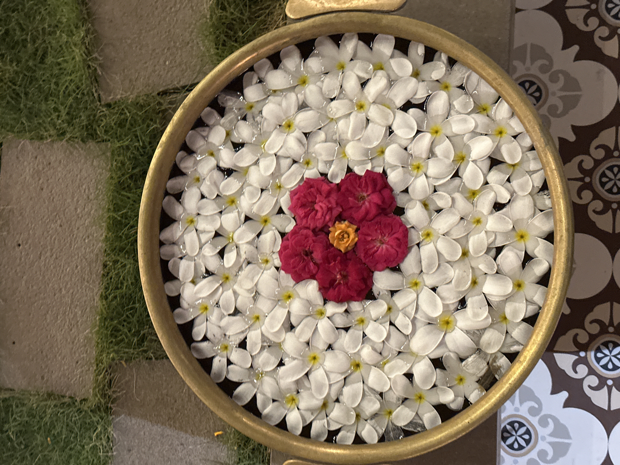 A high-angle shot reveals a circular, gold-colored metal bowl filled with water and floating flowers. Predominantly, the bowl contains numerous white flowers with yellow centers. Positioned in the center of the bowl are several clustered red flowers with a yellow center. The bowl is situated in an outdoor setting. To the left of the bowl is a section of grass and paving stones. To the right, there is a patterned tile surface. The location is likely Kalamb, India.