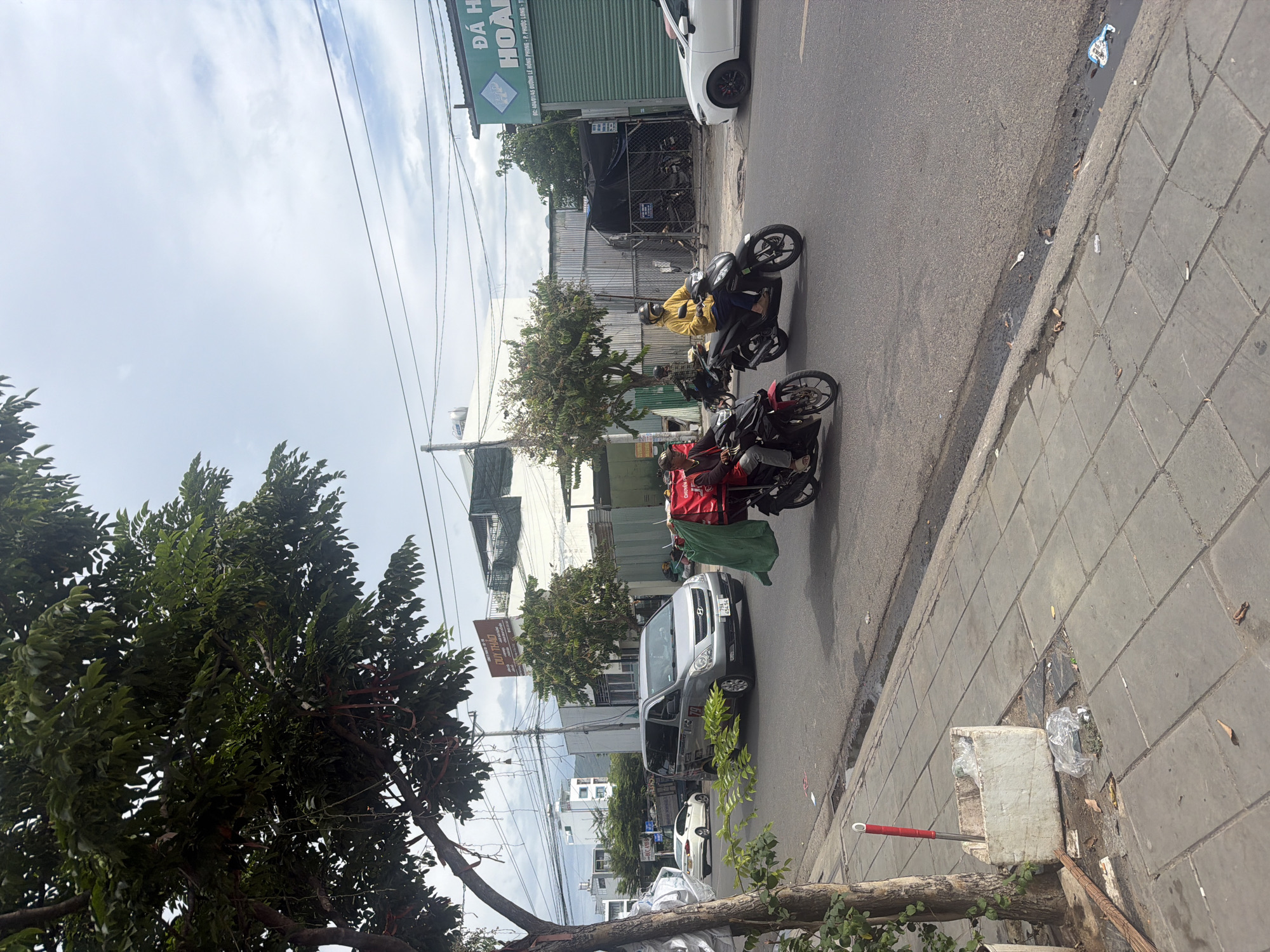 This image depicts a street scene in Nha Trang, Vietnam. On the right side of the road, a person wearing a yellow jacket and helmet is riding a black motorcycle. To their left, another person in a red jacket is riding a black motorcycle. In the distance, a silver van is parked on the side of the road. A white car is also visible further down the street. Overhead, power lines crisscross the sky. The road is paved, and a sidewalk runs along the left side of the image, featuring a white planter and some scattered debris. Trees with dense green foliage line the sidewalk. Buildings and construction can be seen in the background. The sky is partly cloudy.
