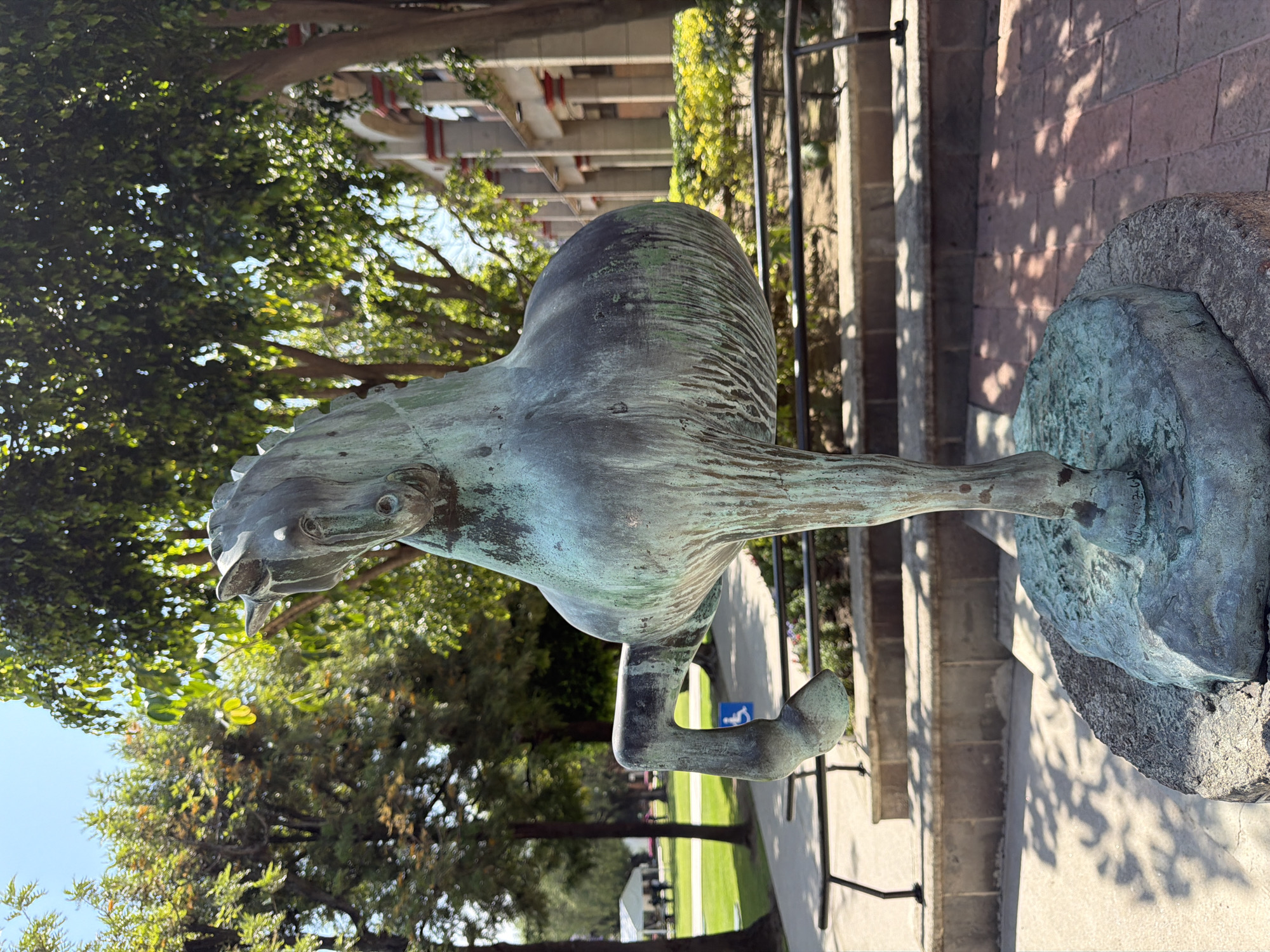 The media file presents a weathered bronze sculpture of a stylized animal, possibly a mythological horse or griffin, set outdoors. The sculpture, exhibiting a greenish-blue patina, stands on a rough stone base. Its head is tilted upwards, featuring defined eyes and a prominent snout or beak, with one foreleg slightly raised.
In the background, vibrant green trees with sunlight filtering through their leaves create dappled light and shadows, set against a clear blue sky. To the right, a brick wall is visible, also marked by strong shadows, alongside a black metal railing and concrete steps descending. Further in the distance, architectural structures with horizontal beams can be seen, along with more trees and a grassy area.
A notable detail in the distant background is a blue sign featuring a white wheelchair symbol, indicating accessibility. The scene is captured under bright daylight conditions, suggesting a sunny morning or afternoon. The overall setting, combining an ancient-looking sculpture with natural elements and architecture in San Andrés Cholula, Mexico, implies a cultural or public space like a park or historical grounds. No specific activity is taking place, focusing instead on the static sculpture and its surroundings.