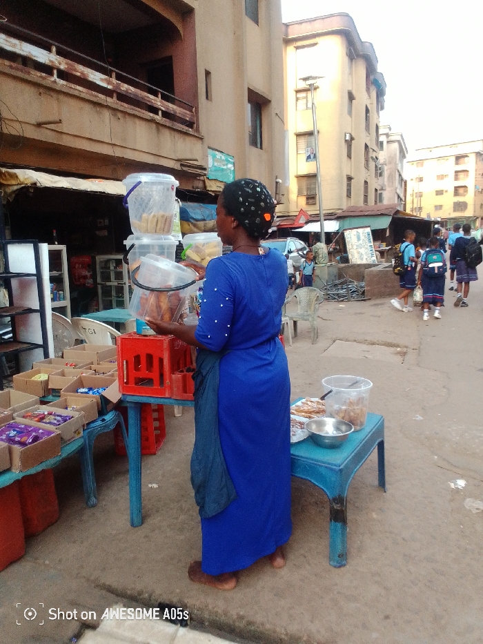 A woman in a bright blue dress stands by two tables on a street corner. She's stocking her stand with clear plastic tubs. One table holds boxes of snacks, while the other displays a metal bowl and a plastic container. Schoolchildren in uniform walk past in the background. The buildings are old, and a street lamp stands tall nearby. A small advertisement sign is seen on a building behind her. The phone's brand that shot this photo is visible on the lower left corner.