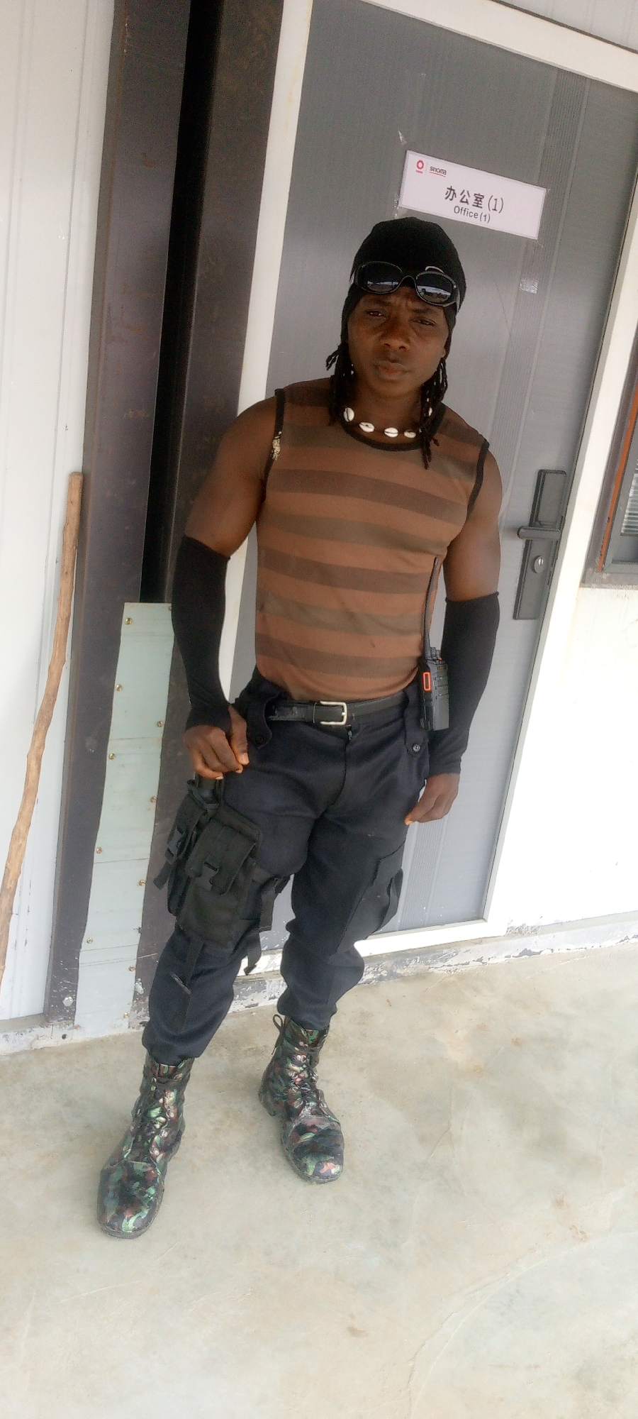 A full-body outdoor shot shows a Black individual standing in front of a gray-framed glass door with a white sign that reads "SINOPEC" and "办公室 (1) Office (1)". The individual is wearing sunglasses, a black bandana, a brown and black striped sleeveless shirt, black arm sleeves, black pants with multiple pockets and straps, and camouflage combat boots. They have a necklace made of shells and a small black radio clipped to their belt. The individual's right hand is resting on their left thigh, and their left hand is slightly bent at the elbow, resting near their hip. The ground appears to be a light-colored concrete surface. A thin wooden stick leans against the door frame to the left of the individual. The environment suggests an urban or semi-urban setting, possibly an industrial or office complex.