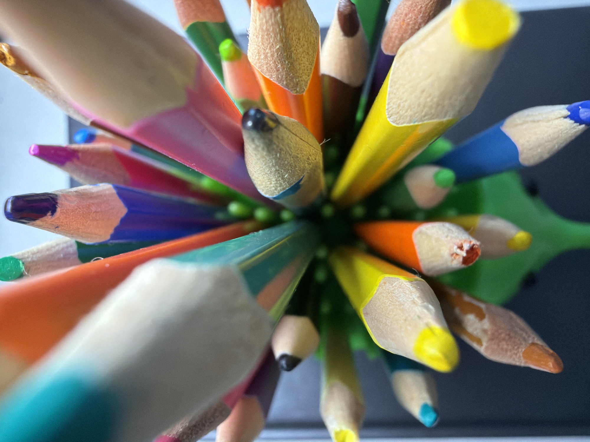 A high-angle, extreme close-up shot depicts a collection of colored pencils densely packed together, likely within a holder. The pencils vary in color, including shades of blue, green, yellow, orange, red, pink, purple, brown, and white. The sharpened tips of the pencils are prominently displayed. The pencil holder appears to be green with small, rounded protrusions. The background shows indistinct surfaces that suggest a desk or tabletop with possibly white and gray areas. No people or specific interactions are discernible. The location is not explicitly identifiable as Votkinsk, Russia, based solely on the visual information provided.