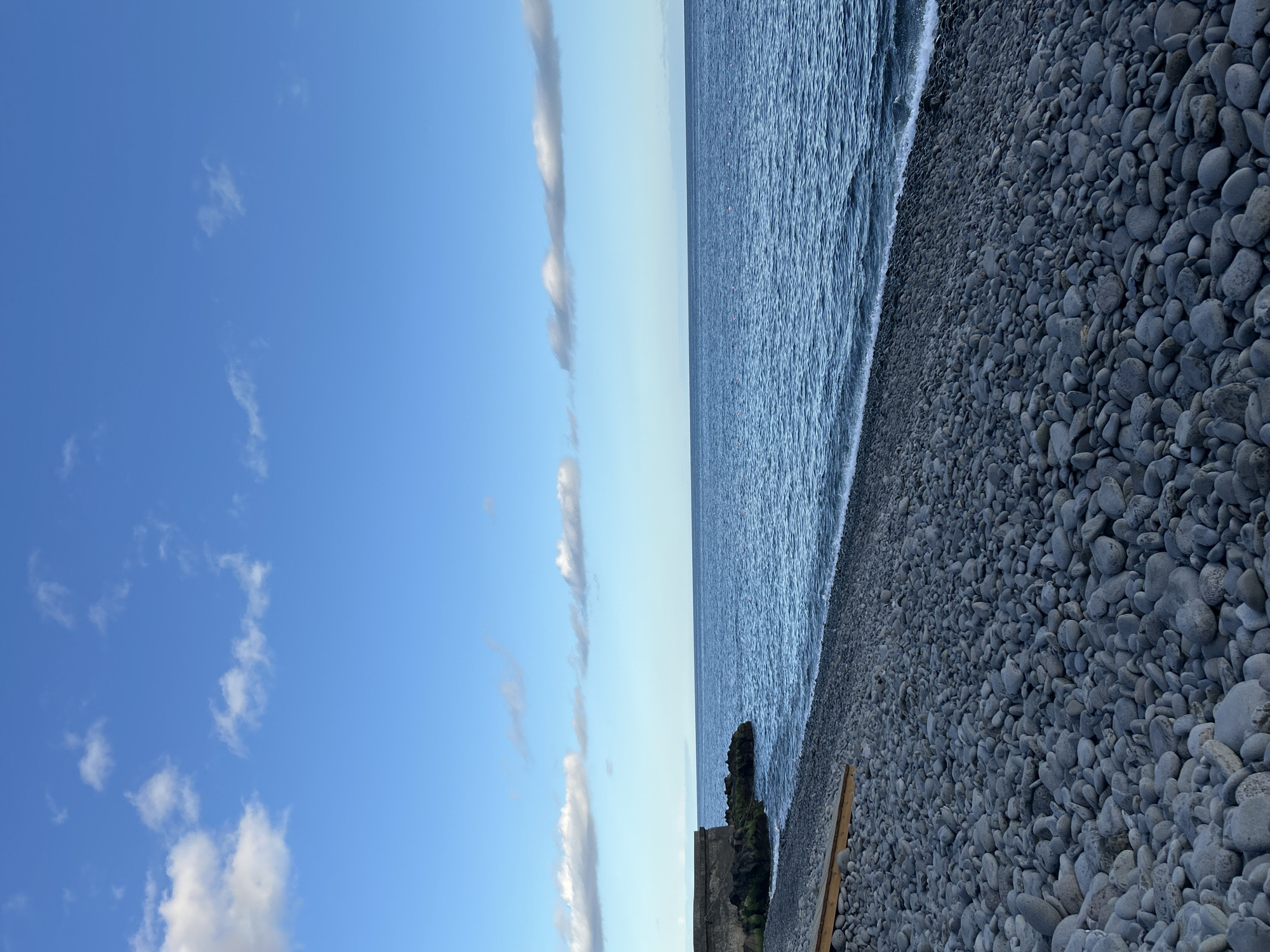 The image depicts a pebble beach adjacent to the sea under a partly cloudy sky. The location is in Câmara de Lobos, Portugal, evidenced by the distinctive grey pebbles commonly found on the shores there.
The left side of the frame is dominated by a blue sky with scattered white clouds. A vertical formation of elongated clouds stretches across the sky.
The sea occupies the central portion of the image, its surface appearing slightly rippled.
To the right of the sea is the pebble beach, densely covered in smooth, grey stones. A portion of a wall structure and some greenery are visible near the shore. A long, possibly wooden, object lies near the shoreline, extending partially into the pebble area. No people are visible in the frame.