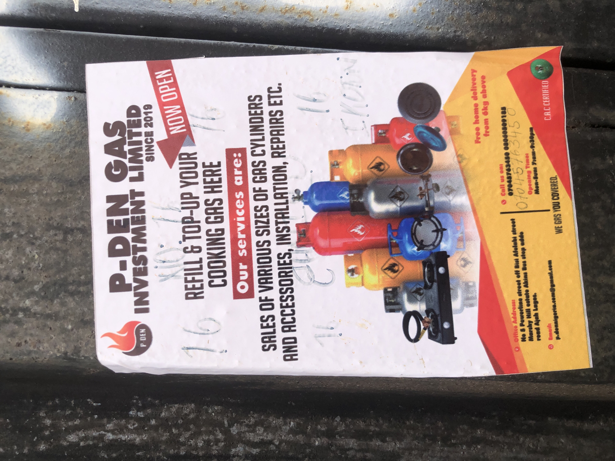 The image shows a flyer for "P-DEN GAS INVESTMENT LIMITED" attached to a dark, textured surface, possibly a metal wall or container. The flyer advertises gas cylinder refills and top-ups, sales, installation, and repairs. It lists an office address at "No 8 Powerline street off Bisi Afolabi street Marshy Hill estate Akins Bus stop addo road Ajah Lagos." Contact information includes a phone number "07048763450 08060309105" and an email address "p.dennigeria.com@gmail.com." The flyer also mentions "Free home delivery from 6kg above" and provides opening hours: "Mon-Sun 7am-9:30pm." The flyer has some handwritten markings and scuffs, indicating it has been posted for some time. The background is out of focus but appears to be a rough, industrial-like environment.