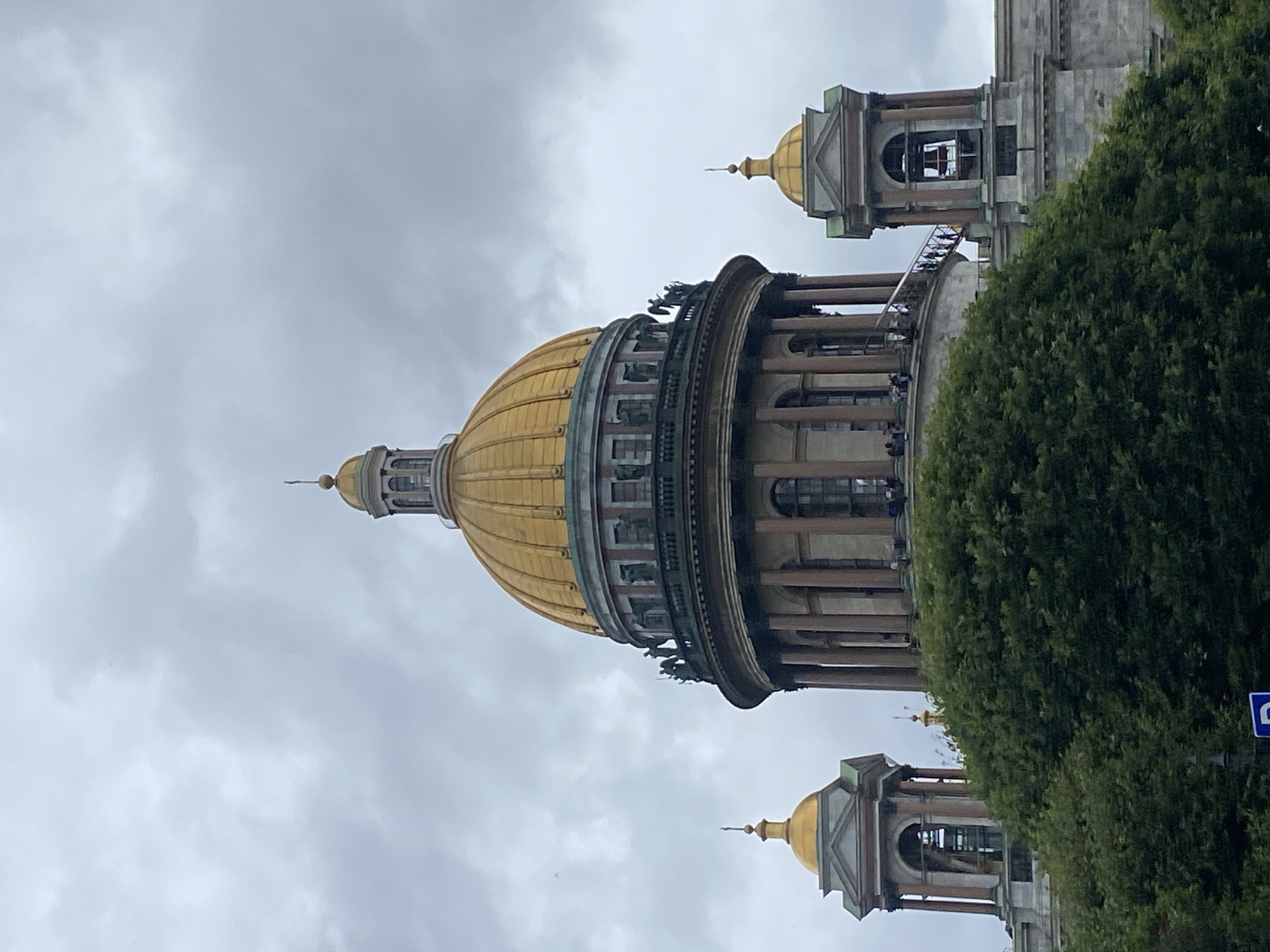 The image shows a partial view of Saint Isaac's Cathedral in Saint Petersburg, Russia, under a cloudy sky. The cathedral features a large, golden dome at its center, surrounded by smaller golden domes on turret-like structures on either side. The main dome is supported by a cylindrical drum with Corinthian columns and arched windows. A balcony, adorned with statues, encircles the drum. A staircase leads up to this balcony from the right side of the main structure, where several people are visible. A lush green tree canopy obscures the lower portion of the cathedral and the ground. The sky is overcast with gray clouds.