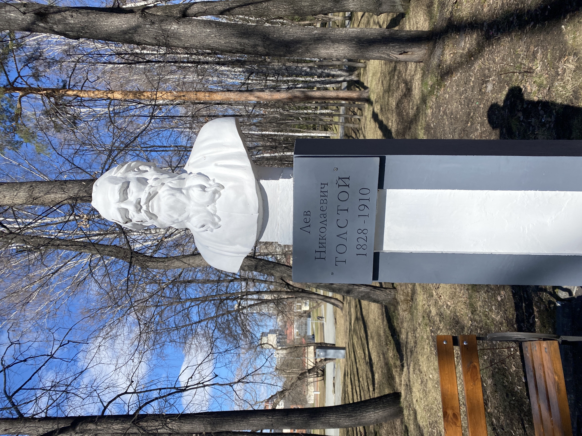 The image shows a monument to Leo Tolstoy in Yekaterinburg, Russia. The bust of Tolstoy is white and placed on a gray pedestal. The monument is located in a park-like setting with trees and grass. There is a bench visible in the foreground. The sky is blue with some clouds, and bare trees are visible in the background.