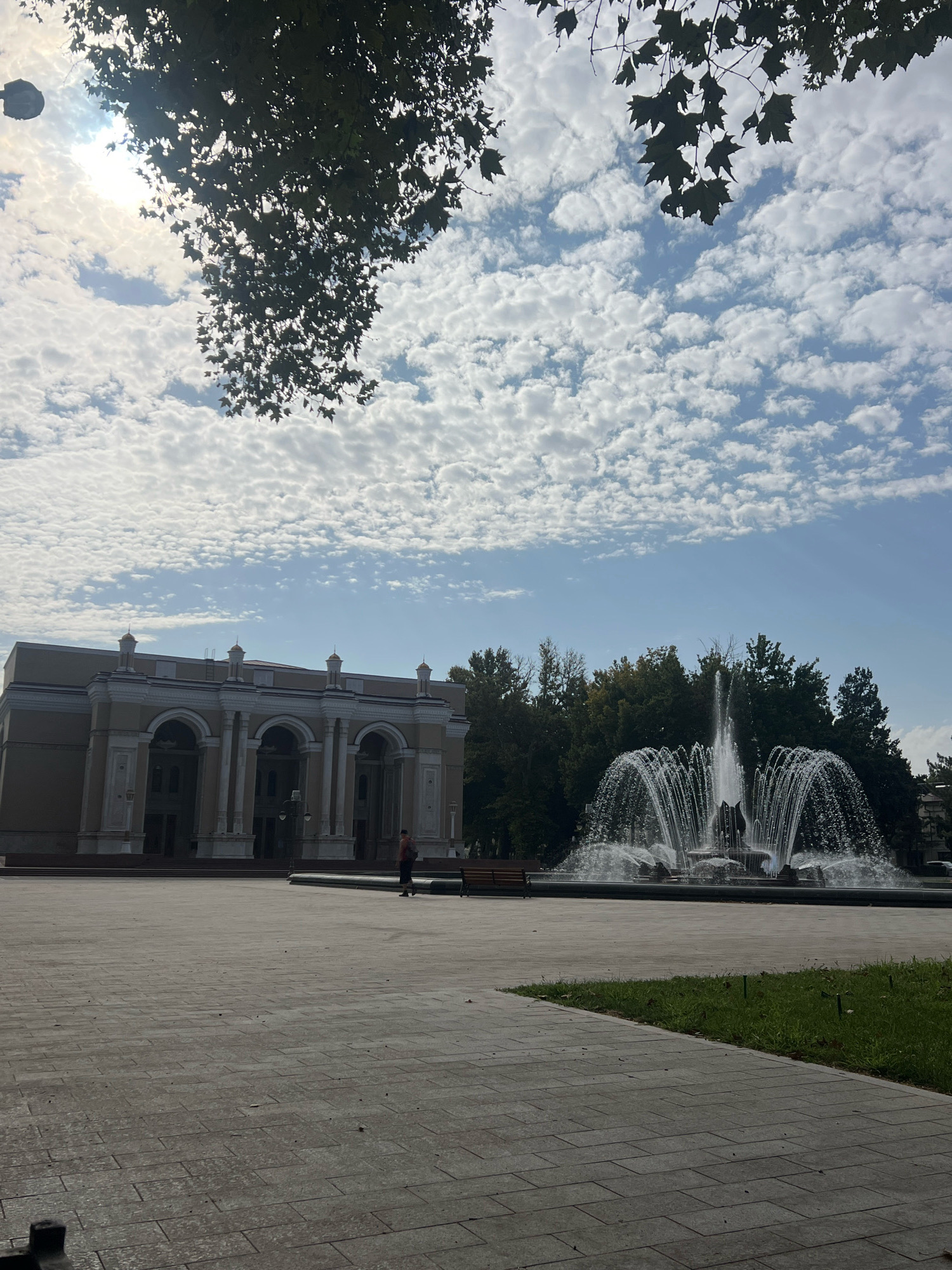A wide-angle outdoor shot captures a public square with a grand building and a large fountain. The building, on the left, features a light-colored facade with multiple arched entrances, ornate columns, and decorative elements. A lone person is walking in front of the building. To the right of the building, a multi-tiered fountain sprays water in a wide arc, set against a backdrop of lush green trees. The square itself is paved with large, rectangular tiles. A strip of grass with sprinklers is visible on the right side of the frame. Overhead, a bright sky is filled with a pattern of small, puffy white clouds, and branches of a leafy tree hang from the top of the frame, partially obscuring the sun. The lighting suggests it is daytime.