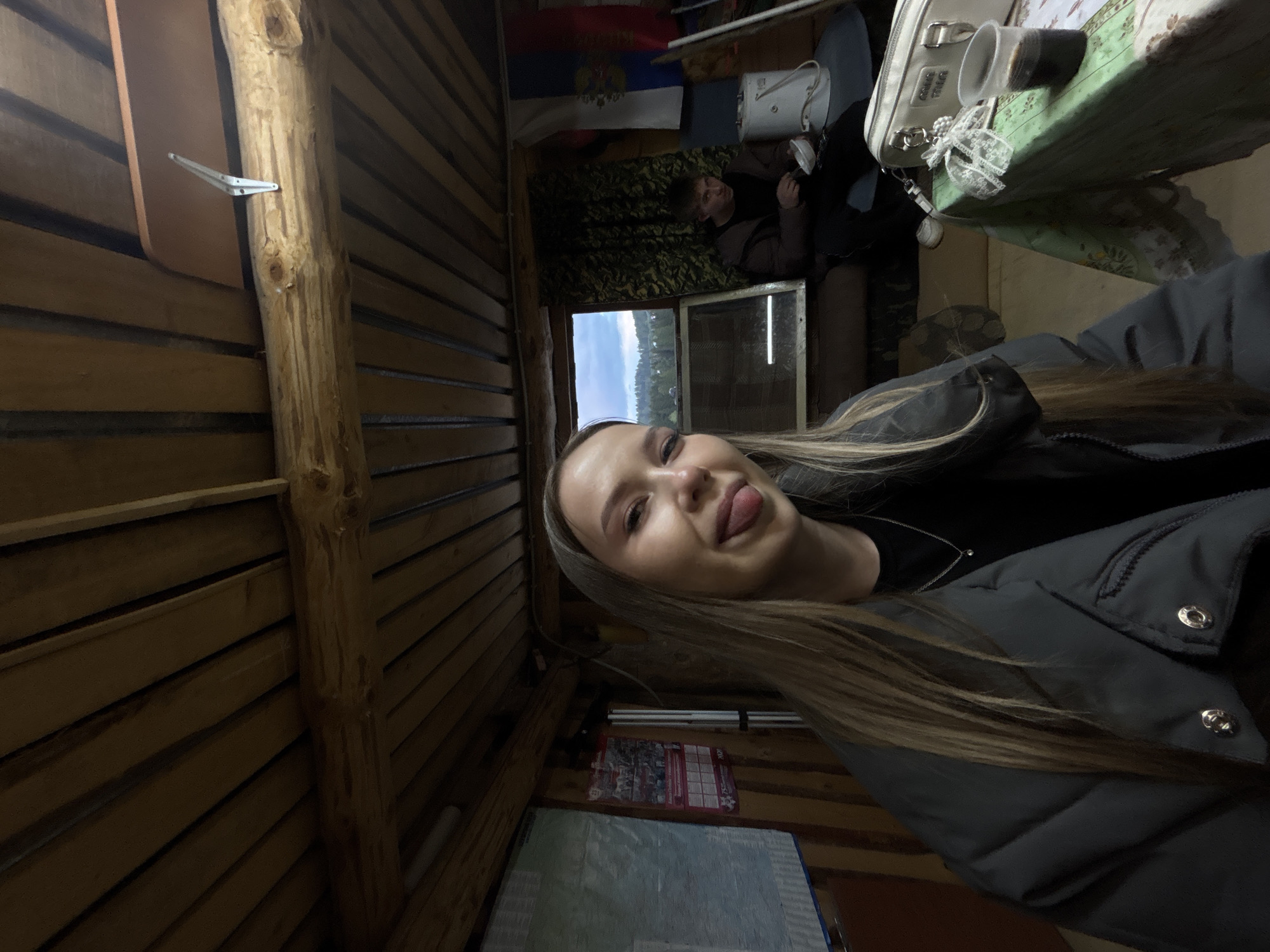 The image shows a woman indoors, sticking her tongue out for the camera. The setting appears to be a cabin or wooden structure, evidenced by the wood paneled walls. In the background, another person is visible sitting on a sofa, with a flag and a window present in the same space. The location is Фоки, Russia.