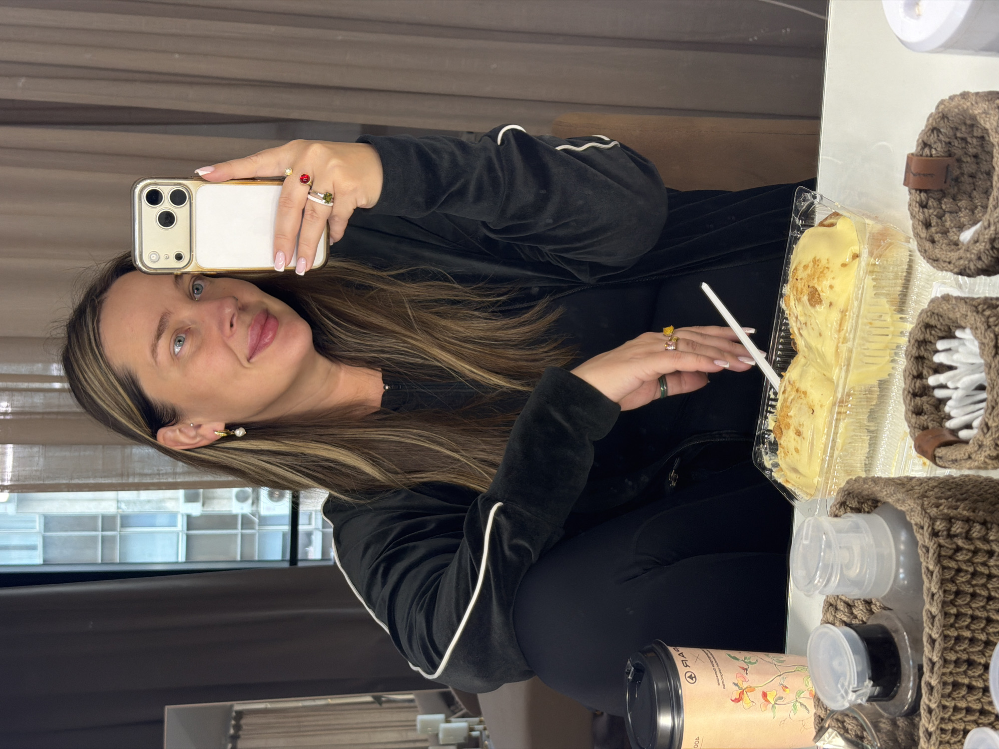 A woman is taking a selfie with a smartphone, holding it in her right hand. She has long, highlighted hair and is wearing a black velvet tracksuit with white piping along the sleeves. Her nails are painted with a French manicure. She is wearing several rings. The background is a neutral tone. The location in the city of Moscow, Russia, cannot be ascertained from the image.
On the right side of the frame, there is a clear plastic container with what appears to be a sweet pastry and a white plastic fork. Several crocheted brown containers holding items like cotton swabs and bottles are present, along with a to-go coffee cup.
