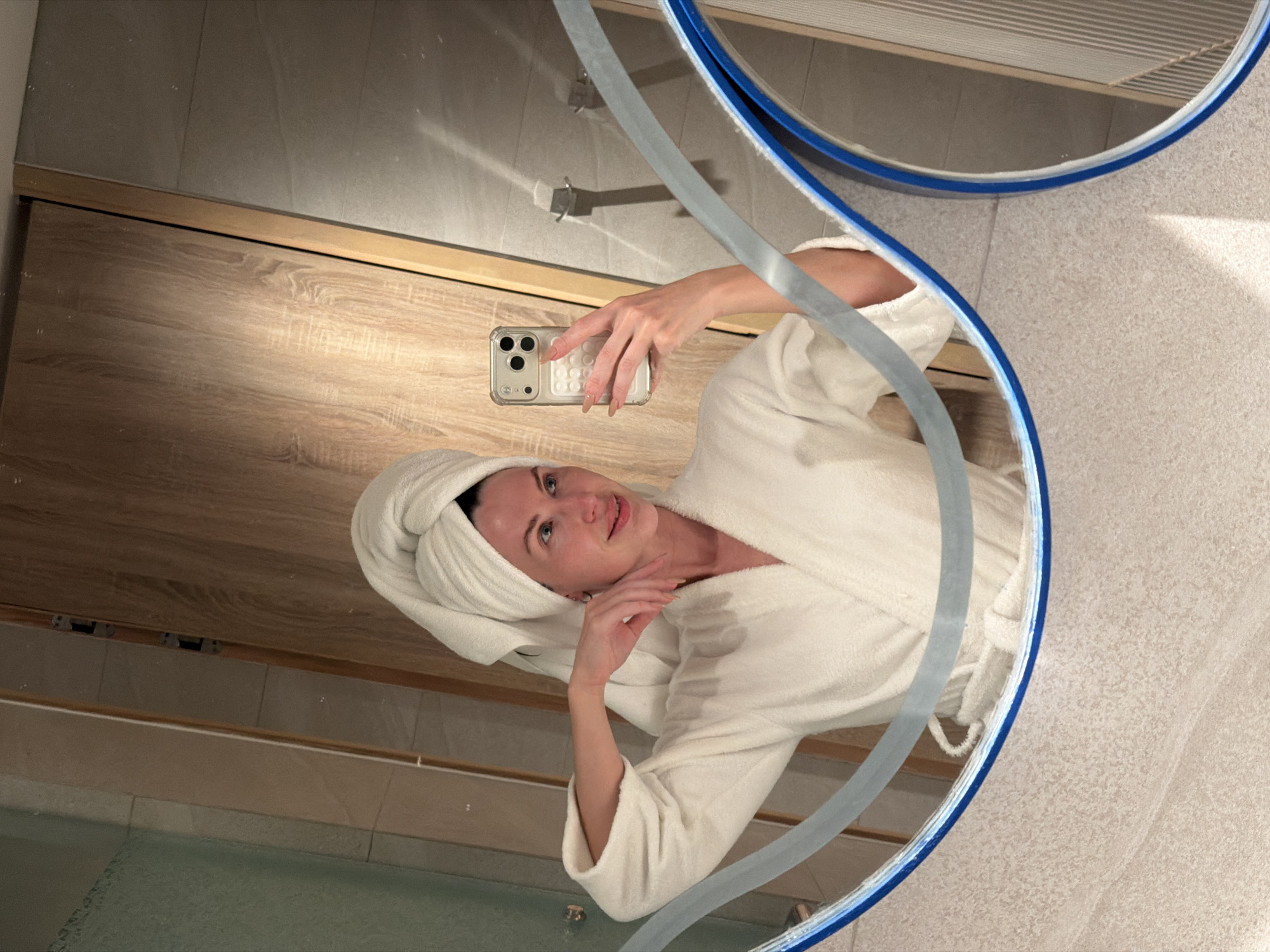 In a bathroom in Dong Ninh Hoa Ward, Vietnam, a woman is captured in a mirror selfie. She is wearing a white terrycloth bathrobe and has a white towel wrapped around her head. With her right hand, she holds a light-colored smartphone with a clear, textured case, aiming it towards her reflection. Her left arm is bent, with her hand positioned near her chin. She looks towards the camera with a subtle smile; her eyes are light-colored, and her eyebrows are well-defined. The reflected background reveals light-toned tiled walls, wooden paneling, and a glass shower enclosure. Two silver wall hooks are visible on the tiled surface. The mirror features decorative wavy light-blue and grey lines across its lower portion and has a blue-rimmed edge. A smaller, circular mirror with a blue rim is also visible in the bottom right of the reflection.