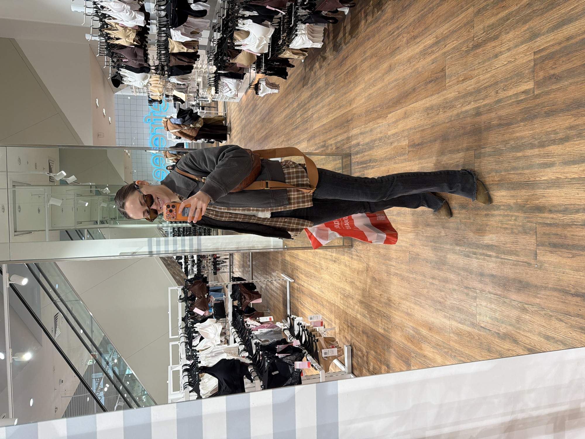 A woman stands in front of a mirror, capturing her reflection with her cell phone. She is clad in dark jeans, a gray sweatshirt, a brown plaid flannel shirt, and tan shoes. A brown leather bag is slung across her shoulder. Her face is partially obscured by sunglasses. The reflection shows a store interior, specifically a section displaying underwear. Rows of bras and underwear hang on racks. In the background, an escalator is visible. The floor has a wood-grain pattern. A shopping bag rests near the woman's feet.