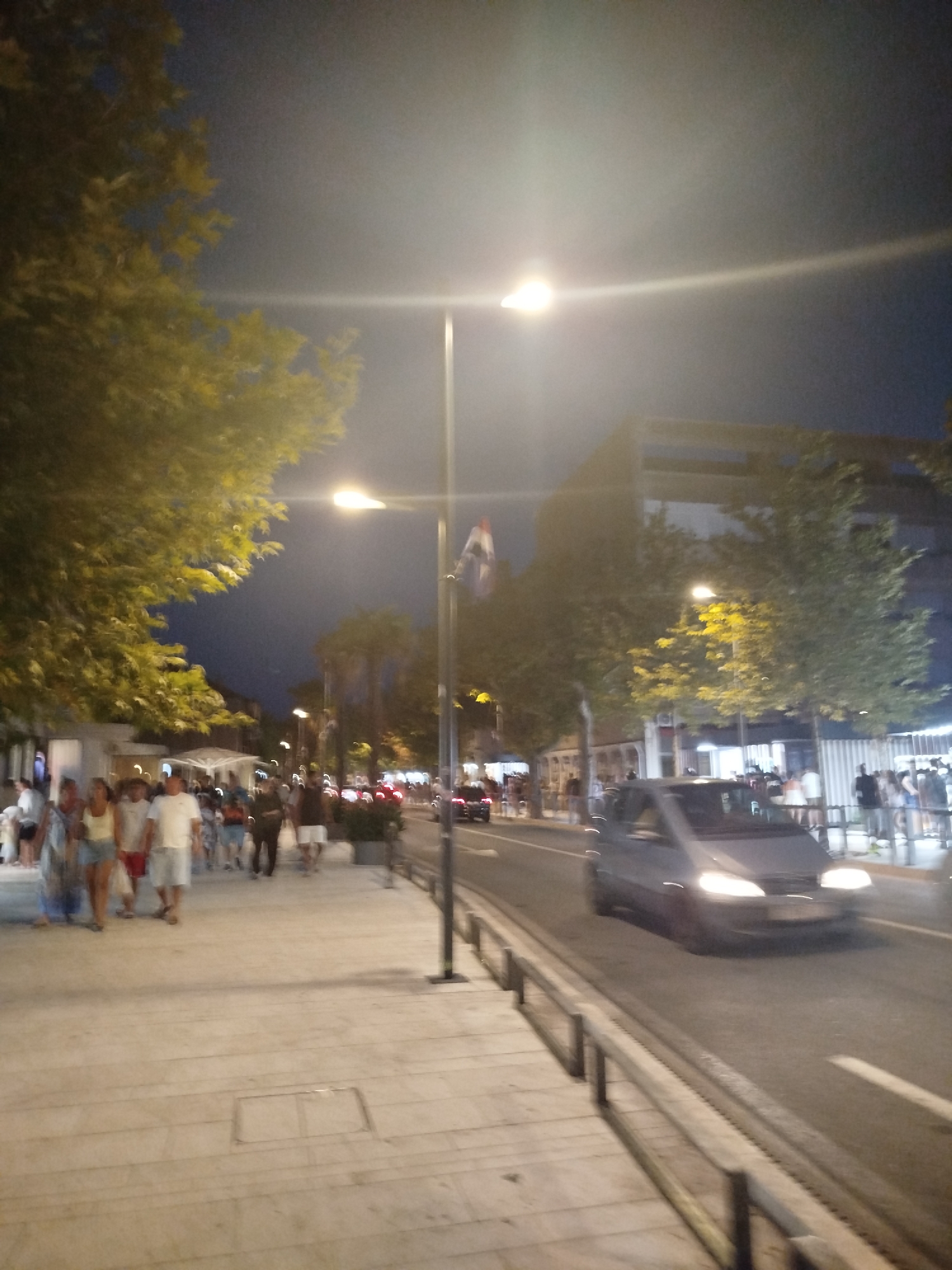 The image captures a nighttime scene on a pedestrian walkway and street in Omiš, Croatia. A group of people are walking along the paved walkway on the left side of the image, illuminated by streetlights. Trees with yellowing leaves line the walkway, casting shadows. Further down the walkway, more people are visible, some in groups, near illuminated buildings and storefronts. A street separates the walkway from the opposite side, where a dark-colored van is driving on the road, its headlights on. Buildings and more trees are visible on the far side of the street, with lights suggesting activity within. The sky is dark, indicating it is nighttime. The overall impression is a lively, urban environment at dusk or night.