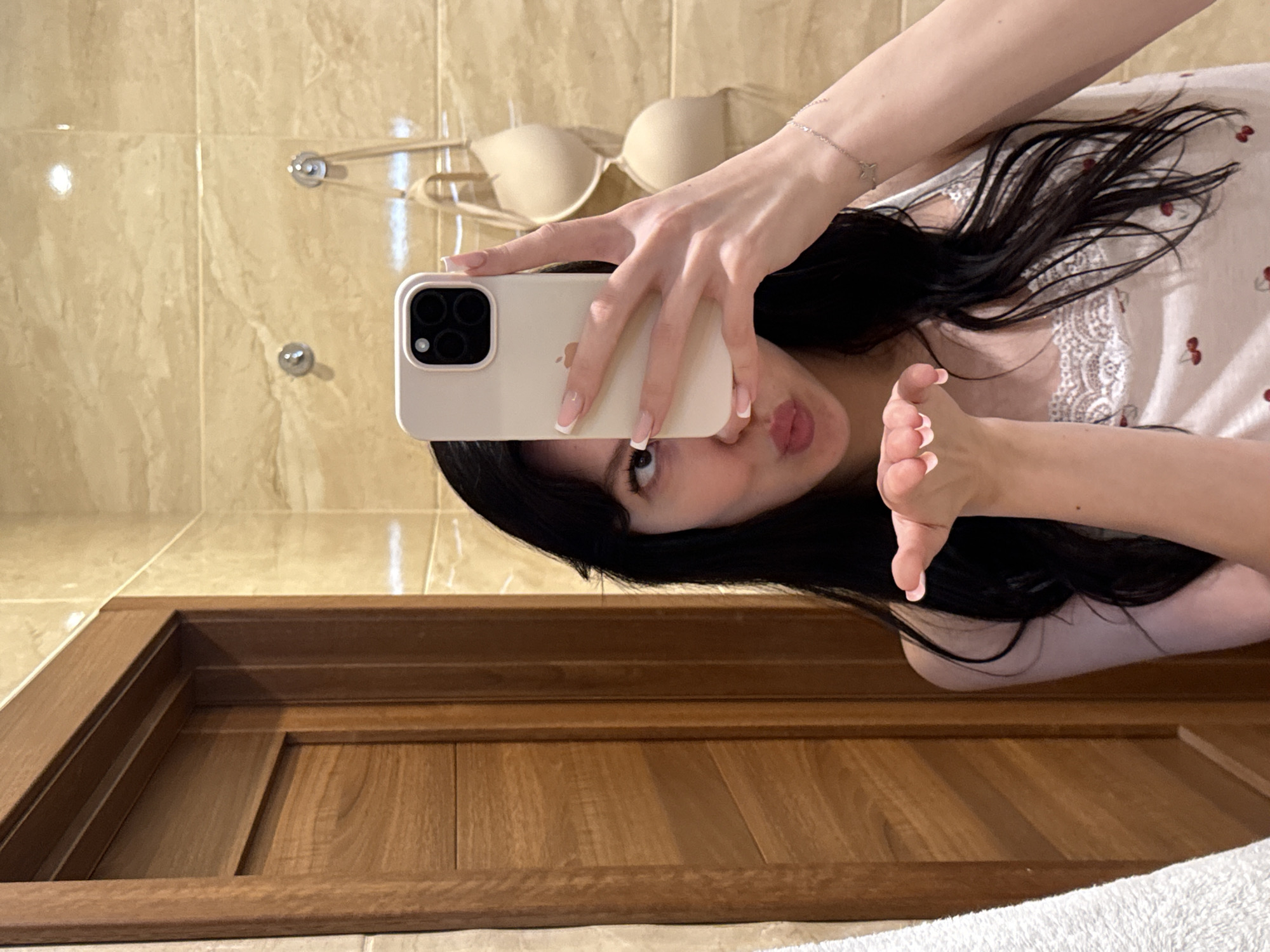 A young woman with long, dark hair takes a mirror selfie in a bathroom. She is wearing a white camisole with a cherry print and lace trim. Her left hand is extended towards the camera as if blowing a kiss. Her right hand holds up a white smartphone, capturing the reflection. In the background, there is a wooden door and beige tiled walls. Two nude-colored bras are hanging on hooks on the wall behind her. The overall environment appears to be a personal bathroom, likely in an apartment or residential building. There is no information in the image to suggest the location is in Khimki, Russia.