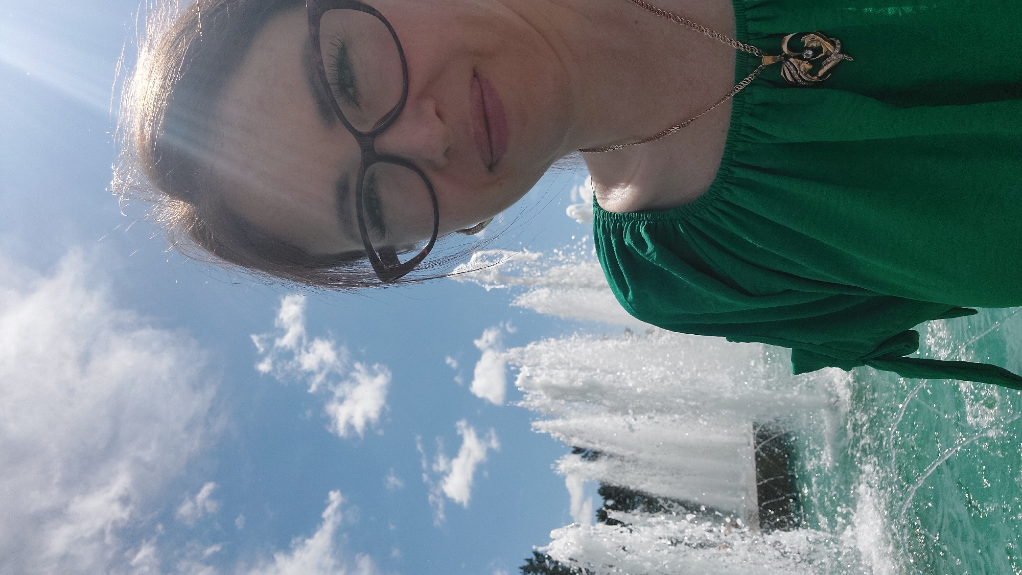 This is a selfie taken outdoors in front of a fountain on a sunny day. The subject is a woman wearing glasses and a green top, with a necklace featuring a heart-shaped pendant. She is smiling and looking towards the camera. In the background, multiple water jets from a fountain are visible, with trees and a blue sky with clouds also present. The scene suggests a public park or recreational area, possibly within the city of Komsomolsk-on-Amur, Russia.