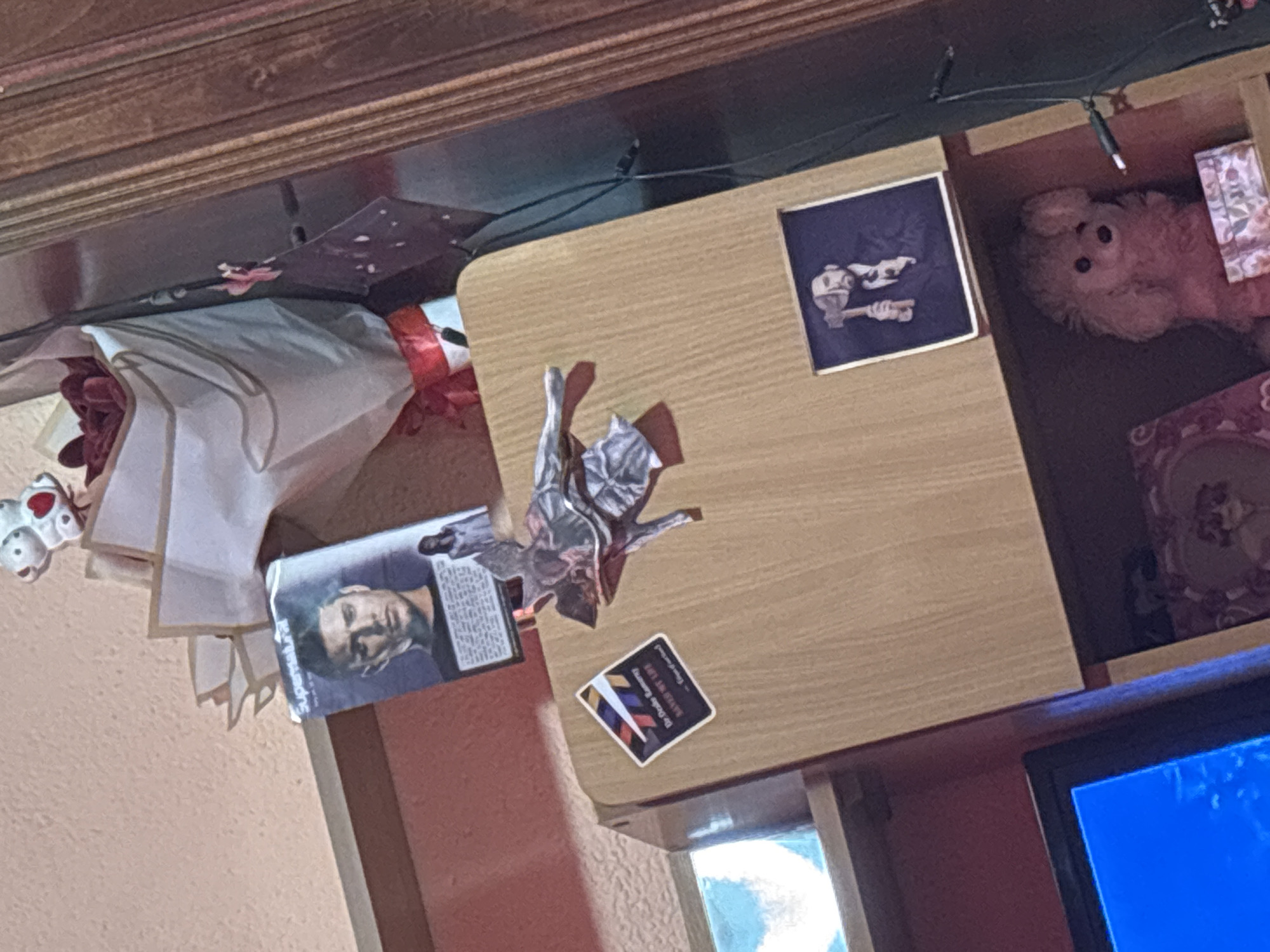 The image depicts a cluttered scene on top of a wooden desk or dresser. Objects present include a white bouquet of flowers, a box with the actor Jensen Ackles from the television show "Supernatural," a folded silver object, a sticker with colored bars, a framed picture of tools, a stuffed animal, and a decorative box. The lower portion of a television screen is visible. The scene appears to be indoors, potentially a bedroom or living area. There is no information to relate this scene to the city of Garmen, Bulgaria.