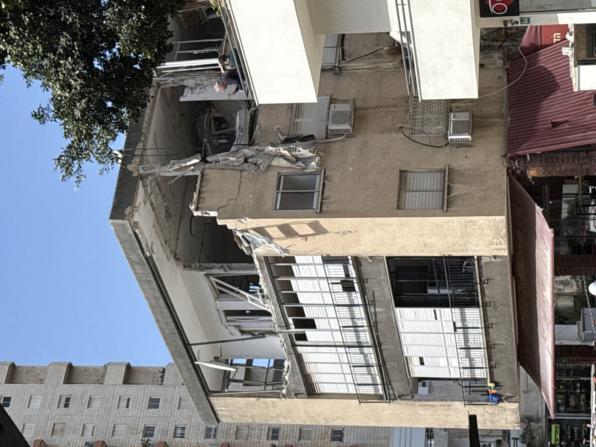 The image shows a damaged building in Ramat Gan, Israel. The building's facade is severely cracked and partially collapsed, with exposed concrete and metal reinforcements. Several windows are broken or missing. A person is visible on the upper floors, surveying the damage. The surrounding environment includes other buildings, trees, and urban infrastructure. A red awning is seen on an adjacent building.