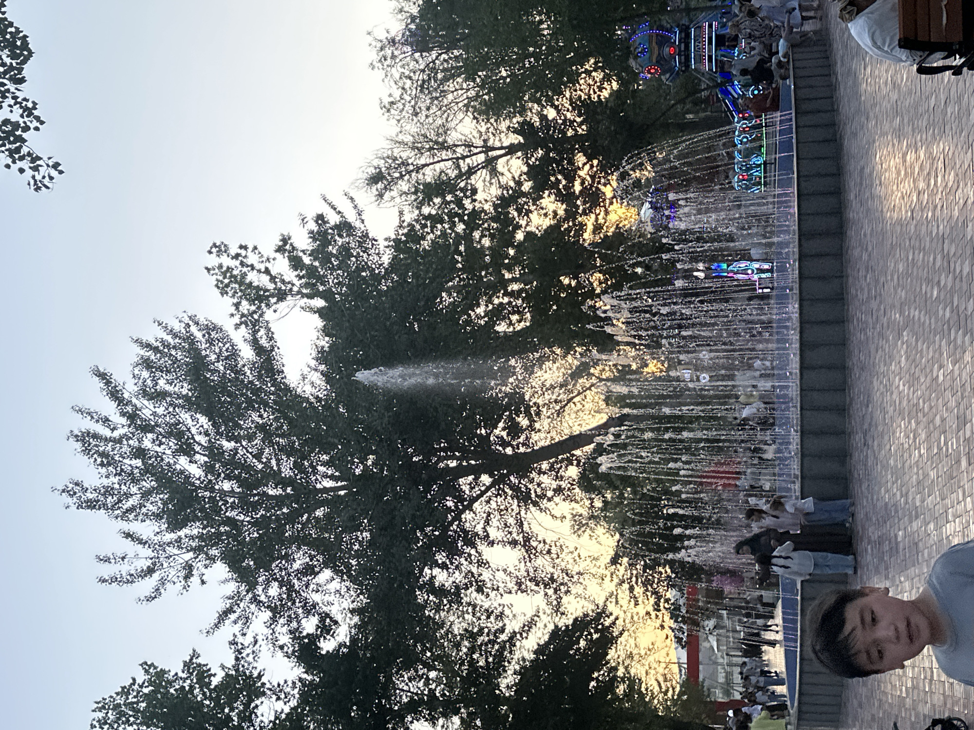 The image depicts a public park area in Manas, Kyrgyzstan, as indicated by the context. In the foreground, a young boy with dark hair is looking towards the camera, his face partially visible. Behind him, a large, multi-tiered fountain is active, with water spraying upwards and outwards, illuminated by colorful lights around its base. The fountain is surrounded by a dark-colored basin, and people are gathered around it, some observing the water, others interacting.
In the middle ground, there are several trees with dense green foliage, some of which are silhouetted against the bright sky. The sky is a pale blue, suggesting either early morning or late evening. There are also hints of a carnival or amusement ride in the background, with bright lights and a colorful structure. A path paved with brick-like material extends through the park. The overall scene suggests a lively public space with recreational activities.