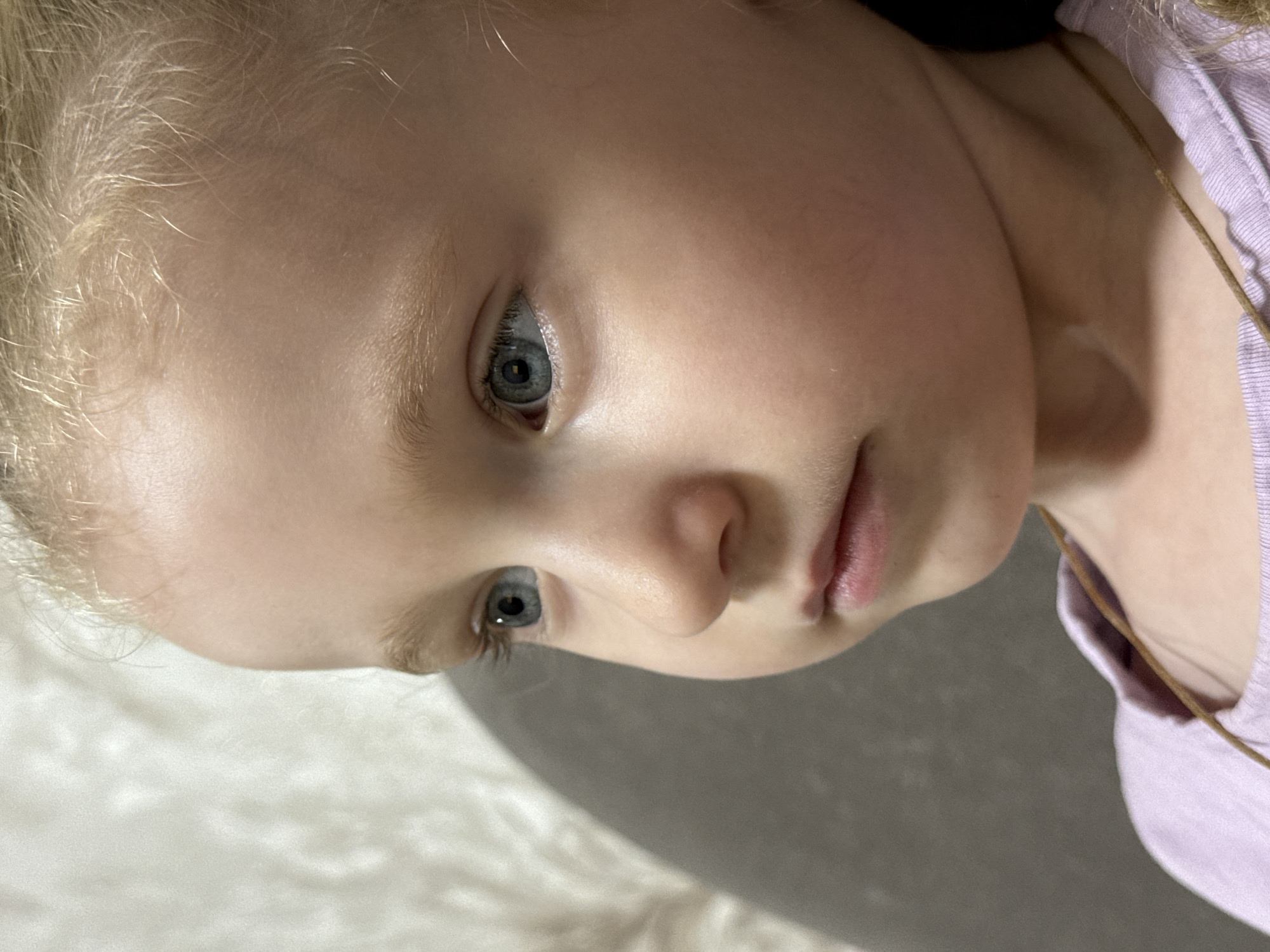 A close-up, profile shot shows a young Caucasian girl with blonde hair and blue eyes. She is looking to the left of the frame, her gaze fixed on something unseen. Her skin is fair with rosy cheeks, and her lips are slightly parted. She is wearing a pale lavender t-shirt. A thin, brown string necklace is visible against her neck and shirt. The background is softly blurred, with a textured wall on the left and a darker, indistinct area on the right, possibly a piece of furniture or clothing. The overall impression is one of quiet contemplation or observation. This image was taken in Chaikovsky, Russia.