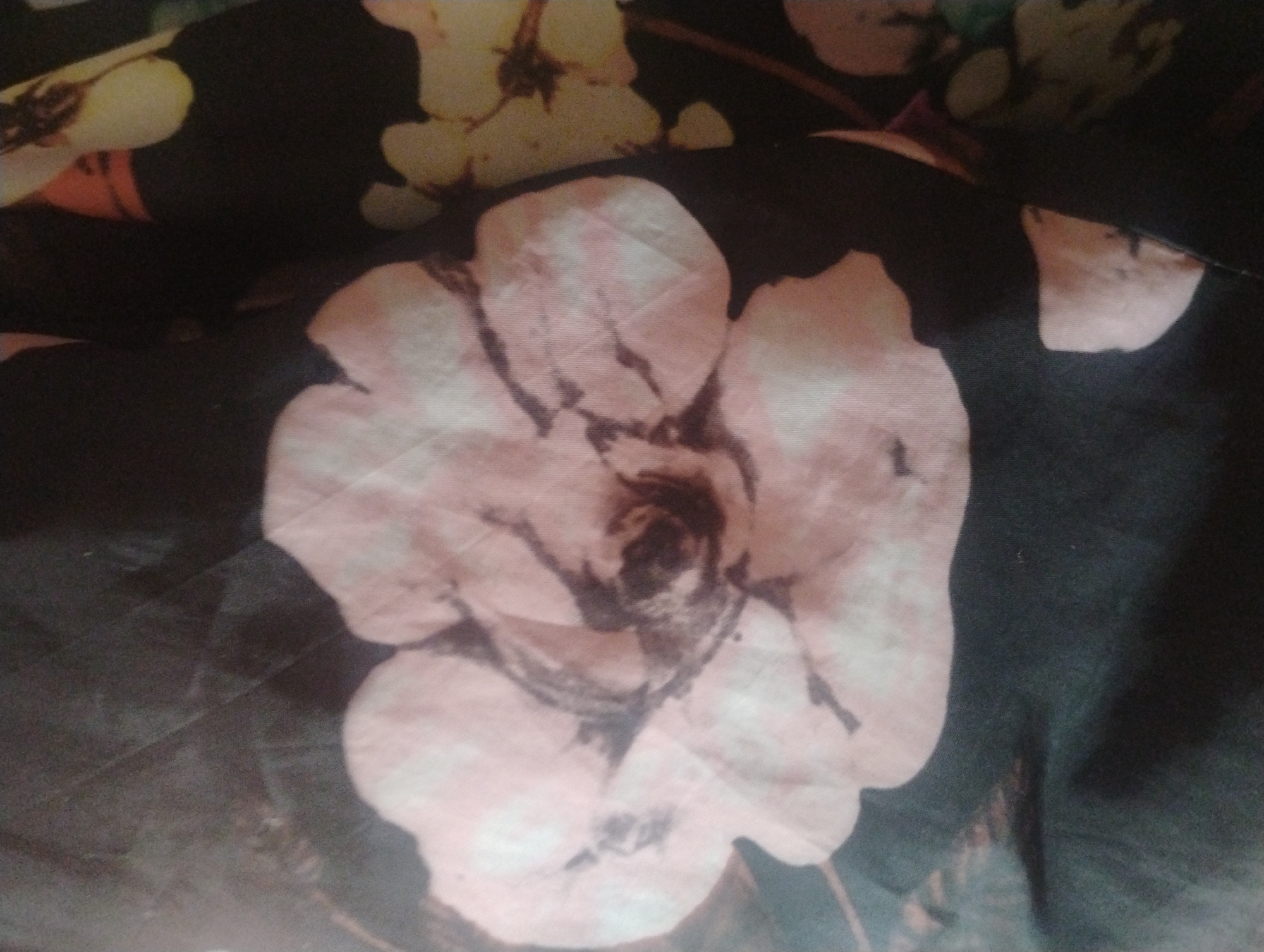 A close-up shot of a dark fabric with a floral pattern. The flowers are rendered in shades of pink, yellow, and brown, with some petals appearing translucent. The pattern includes large, stylized flowers with visible veins and centers. The fabric is textured, with some areas appearing slightly rumpled or creased, suggesting it may be a piece of clothing or upholstery. No people or specific environmental details are visible, and the location in Rumyantsevo, Bulgaria cannot be determined from the image.