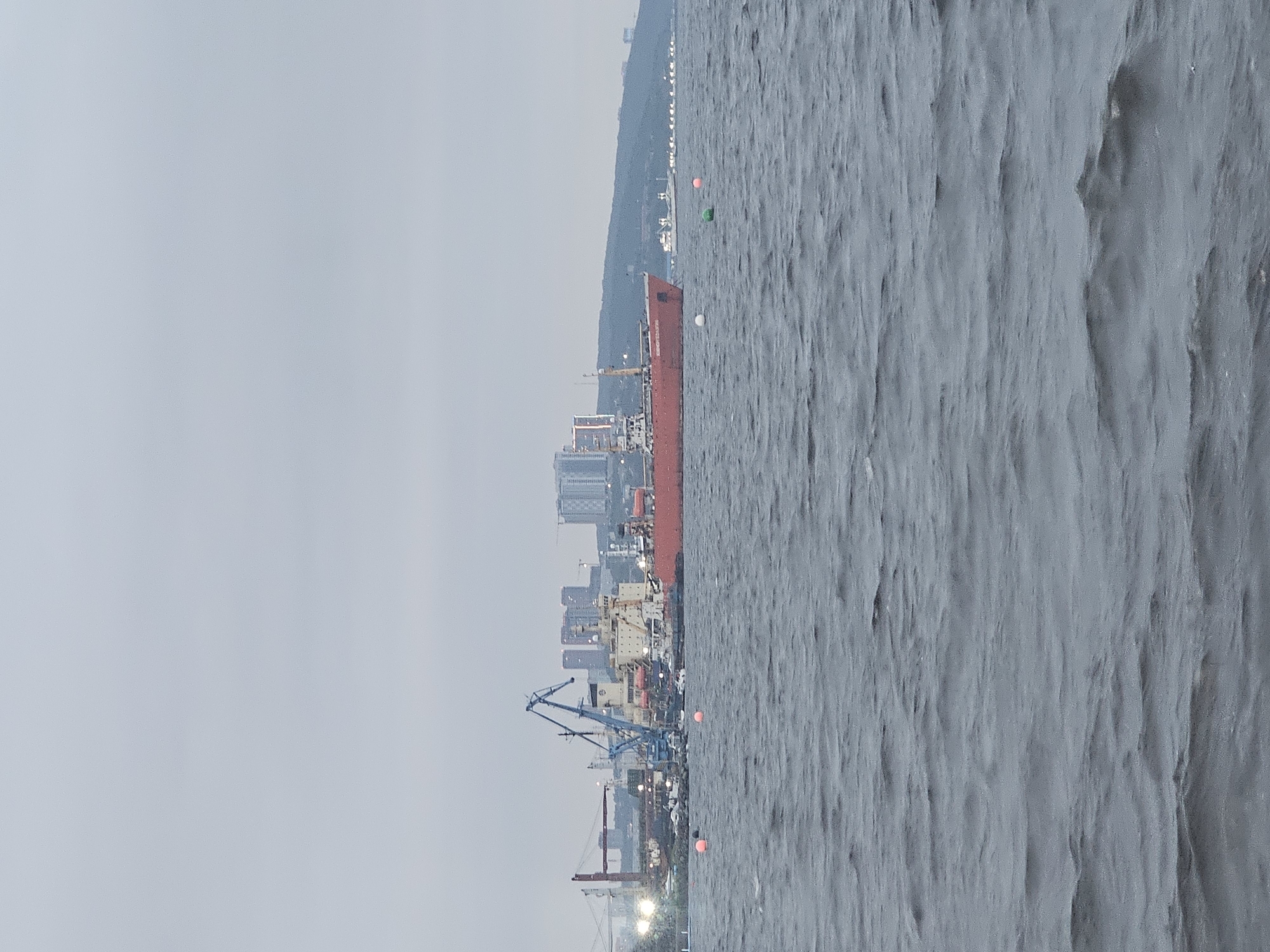 The image depicts a view of the Golden Horn Bay in Vladivostok, Russia, on an overcast day. The foreground is dominated by choppy, dark grey water. In the middle ground, several large ships are docked at a port. The most prominent vessel is a long, red cargo ship with "KOBE EXPRESS" written on its side, suggesting it is a container ship. Beside it, a lighter-colored ship, possibly a tanker or research vessel, is visible. Several cranes, one of which is a tall, blue harbor crane, are positioned on the dock, indicating ongoing port operations. In the background, the cityscape of Vladivostok rises on a hill, with several modern buildings and some older structures. A red suspension bridge, possibly the Russky Bridge or Golden Bridge, is visible on the left side of the frame, with its lights illuminated. Several buoys, two orange and one green, are floating in the water, marking a channel or hazard. The overall atmosphere is muted due to the cloudy weather.