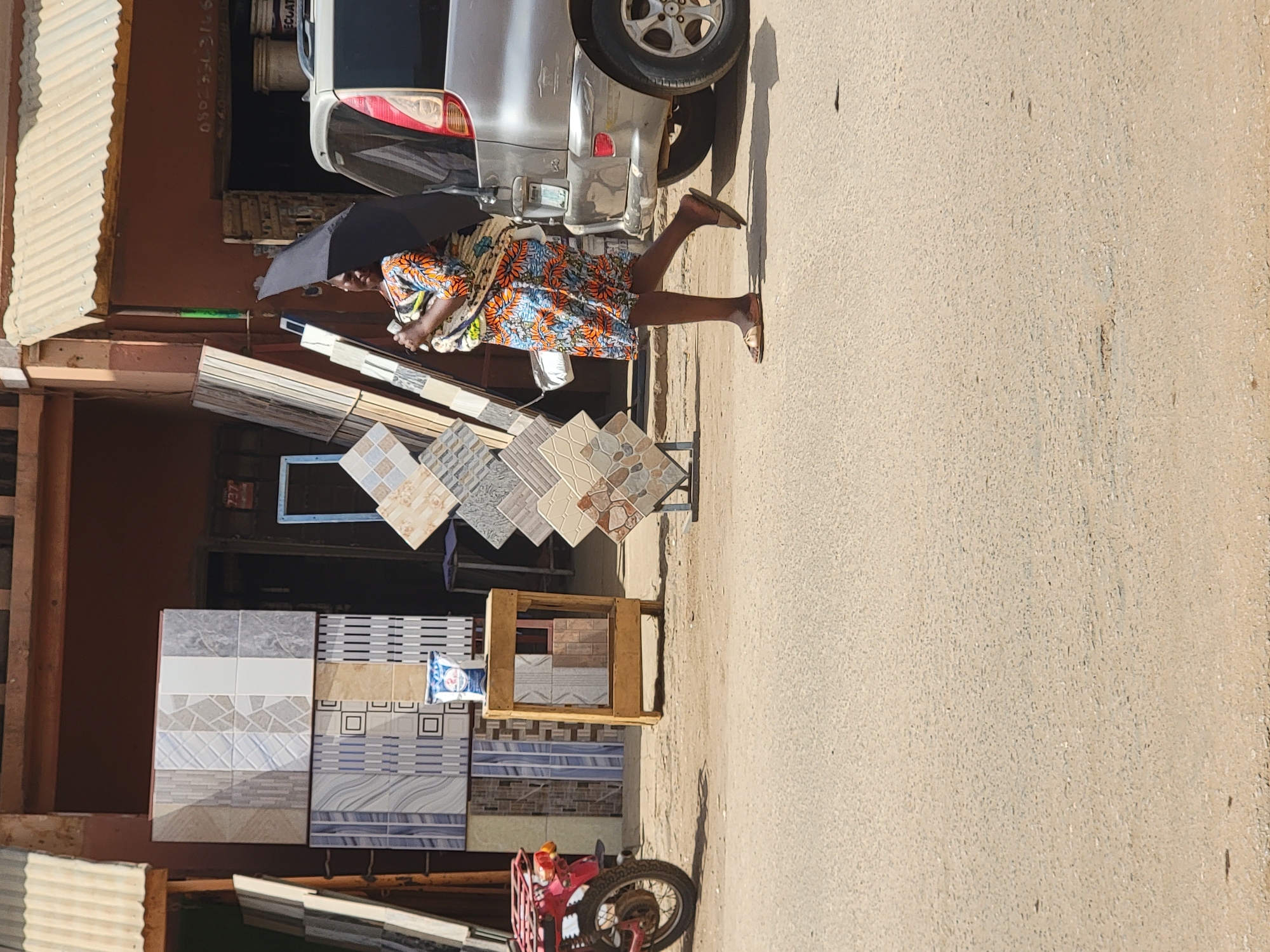 An outdoor scene in Maiburiji, Nigeria, depicts a bustling street or market area under bright daylight. In the center, a person, likely a woman with dark skin, stands wearing a vibrant orange, blue, and white patterned dress and flip-flops. She holds a black umbrella overhead, presumably for shade from the sunny weather, which is indicated by distinct shadows cast by her and a nearby vehicle.
The foreground and midground are dominated by an extensive display of decorative floor and wall tiles. These tiles, showcasing various patterns, colors, and textures (including geometric, mosaic, and plain designs), are arranged on metal stands, a wooden frame, and directly on the ground, suggesting a tile vendor's setup.
Behind the person, a silver/grey car is partially visible, with its rear wheel and bumper prominent. Further back, a building with a reddish-brown wall features visible white text, including what appears to be a phone number: "08064930164". Stacks of other building materials and white buckets, one with "EQUAT" visible, are also seen near the building and car. In the lower right of the frame, part of a red motorcycle or scooter with a front basket is visible. The ground is a light-colored, dusty surface. The image captures an everyday commercial interaction or display in an African street setting.