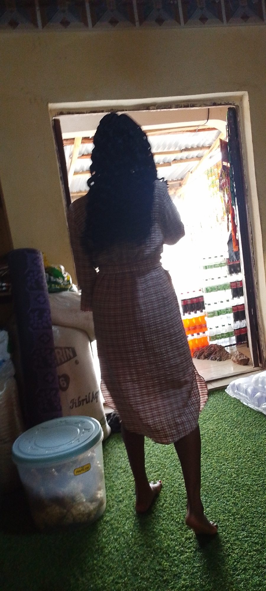 A person with dark hair stands in a doorway. They are facing a bright storefront in Adankolo, Nigeria. The person wears a plaid dress, and their bare feet are visible. To their left, inside the room, are stacked bags, a rolled-up textile, and a plastic container. The storefront visible through the doorway is stocked with drinks.