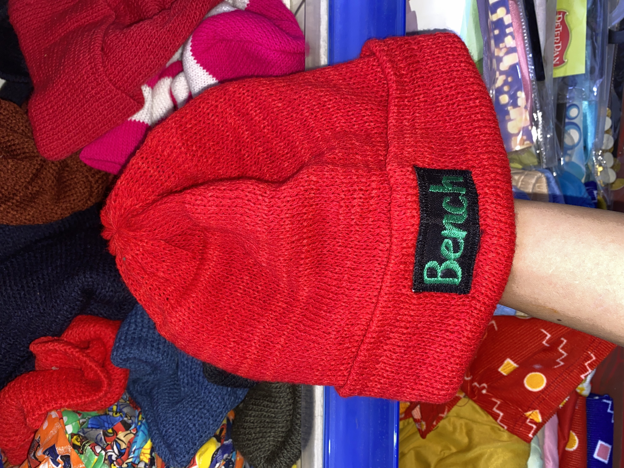 A red knitted beanie with the word "Bench" embroidered in green on a black tag is being held by a hand in the foreground. The beanie is positioned centrally, with its cuff folded up. In the background, various other knitted items and fabrics are visible, including a dark blue beanie, a red knitted fabric, and patterned fabrics. The items appear to be stacked or piled on shelves. The overall environment suggests a retail setting or a personal collection of clothing items. The image does not provide specific geographical markers to pinpoint its location within Davao City, Philippines.