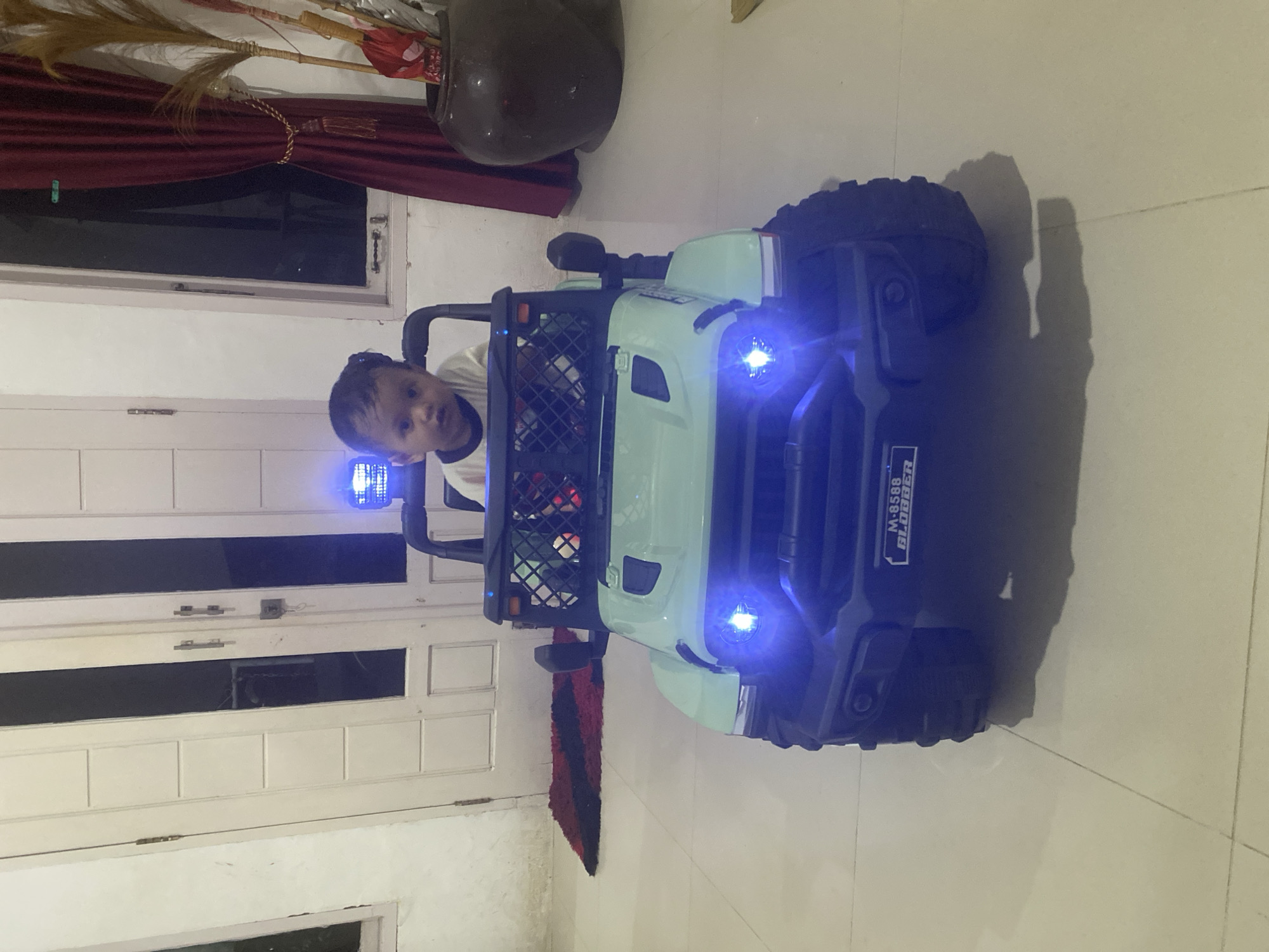 A child is seated in a light green toy jeep-like vehicle, which has functioning headlights that emit a bright blue light. The vehicle is positioned in an indoor space that appears to be a home, judging by the tiled floor and paneled wall with a window. In the background, there is a dark-colored urn or decorative pot. The red drapes suggest the location may be in Lhokseumawe, Indonesia, based on common Indonesian home interior features.
