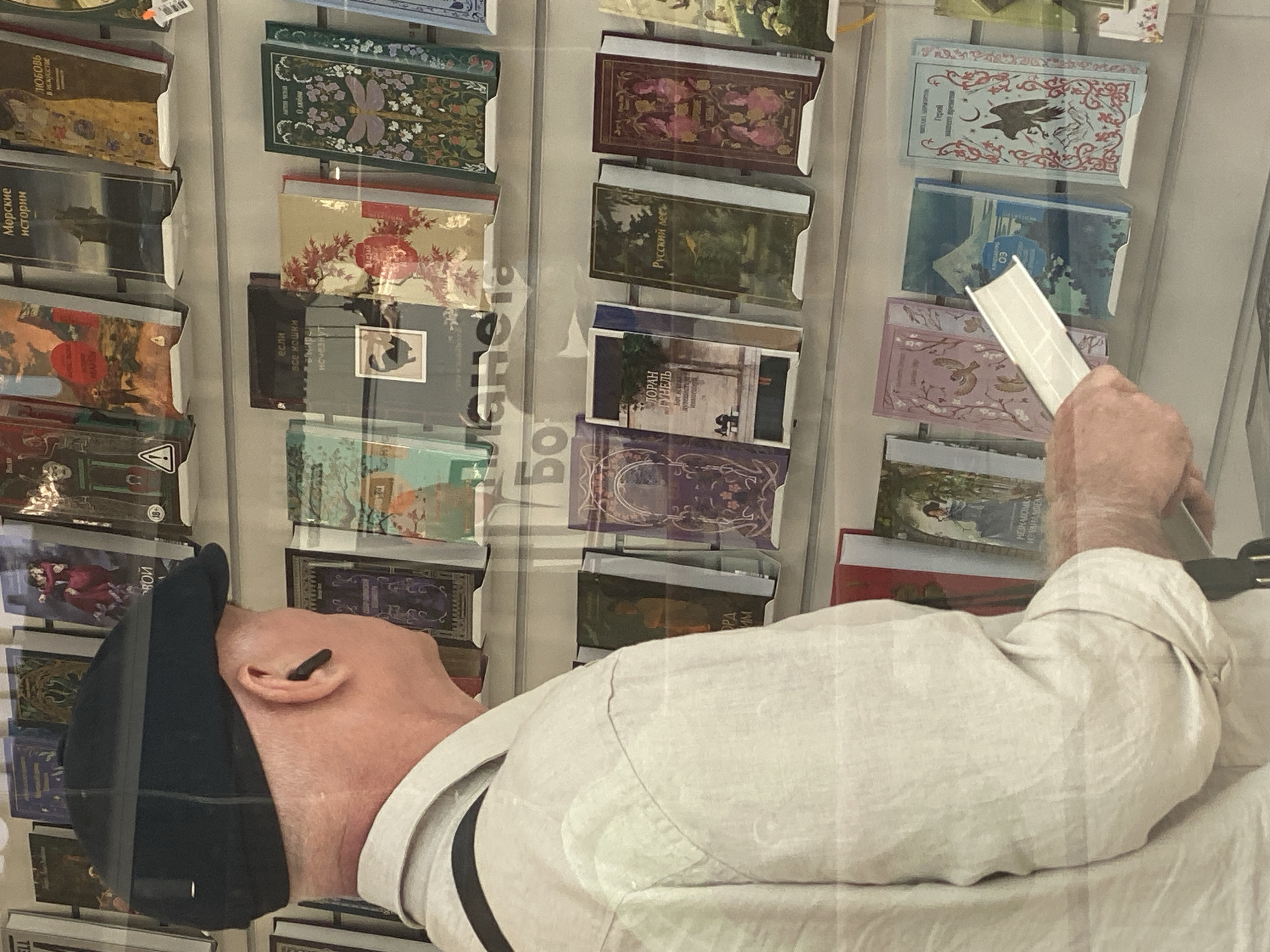 The image shows a man wearing a black flat cap and a light-colored shirt, standing in front of a shelf of books. He has a wireless earbud in his right ear. His left hand is holding a white object, possibly a brochure or a slim book, and his right hand is resting on the shelf. The books on display are mostly hardbound with colorful covers, some featuring illustrations and titles in Russian. The lighting suggests an indoor setting, likely a bookstore or a library. The reflection on the books indicates the image was taken through a glass surface. The city of Novokuznetsk, Russia, is mentioned, but there are no specific visual cues within the image to confirm this location.