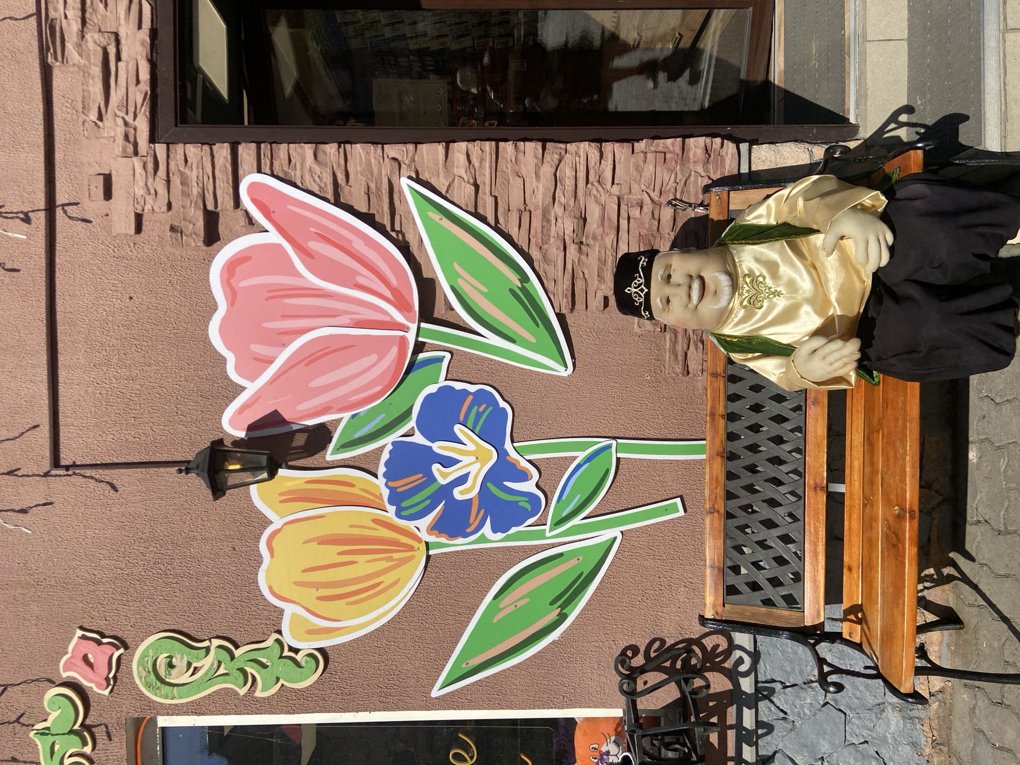 A brown, two-story building is partially visible. Attached to the wall is a large, decorative flower arrangement and the start of a restaurant name in block letters. Adjacent to the flower arrangement is a small, wooden bench with a male mannequin figure sitting upon it. The mannequin is wearing a gold-colored silk shirt with green and gold detailing and a black cap. The manequin appears to be smiling. The bench has a black metal framework. The building appears to be located outdoors in the city of Kazan, Russia.