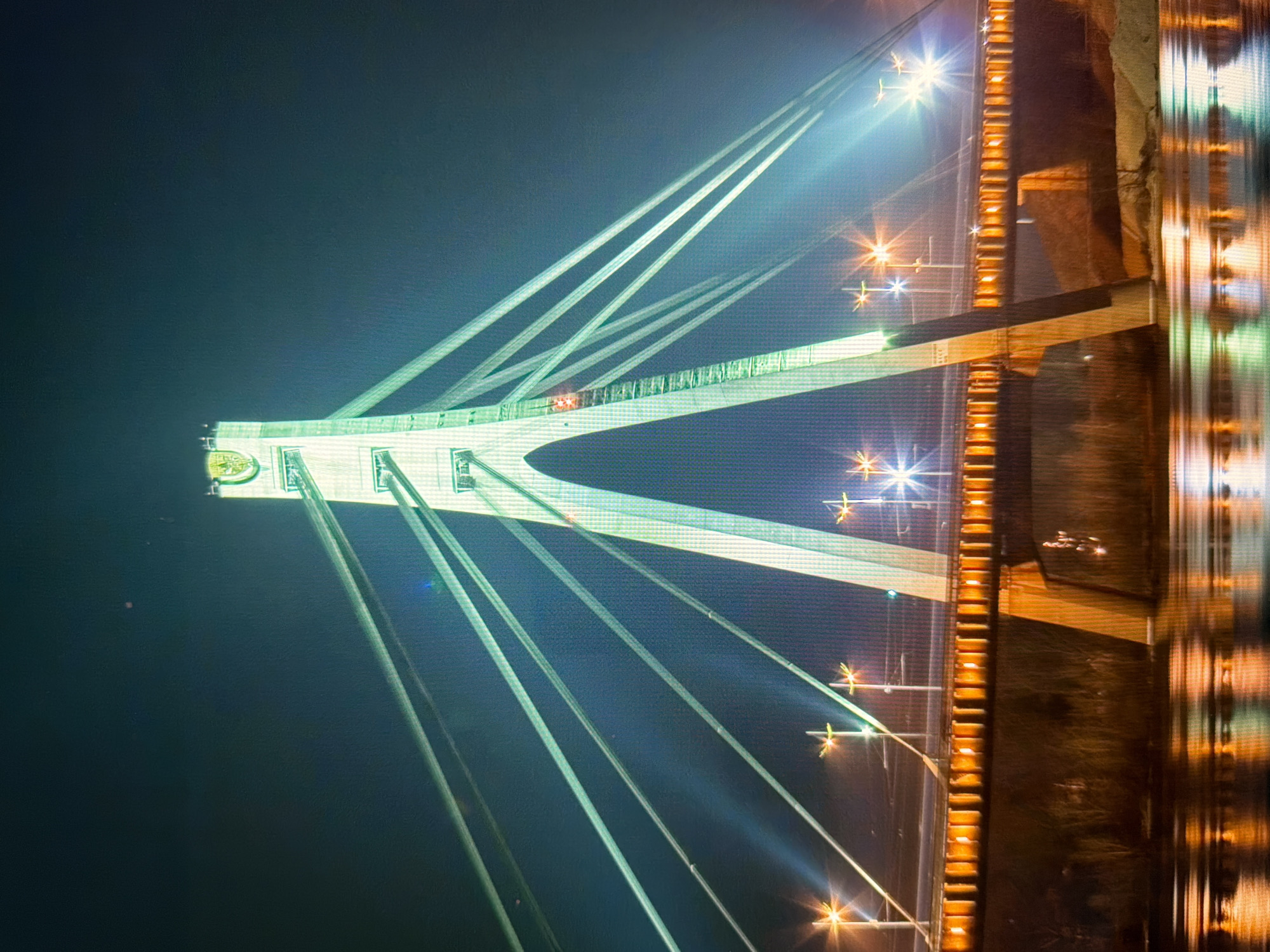 The image shows a nighttime view of the Southern Bridge (Polukrovsky Bridge) in Kyiv, Ukraine. The bridge is a cable-stayed bridge illuminated with green lights on its pylons and white and orange lights along its deck. The reflection of the bridge lights is visible on the dark water of the Dnipro River below. Several streetlights with starburst effects are also visible along the bridge's walkway. The sky is dark and clear, with a few faint lights and a small reddish speck. The bridge's iconic pylon, shaped like a large A-frame, is the central focus of the image, with numerous cables stretching from it to the bridge deck. The banks of the river appear to be lined with sparse vegetation and sand. The overall scene depicts a well-lit urban infrastructure at night.